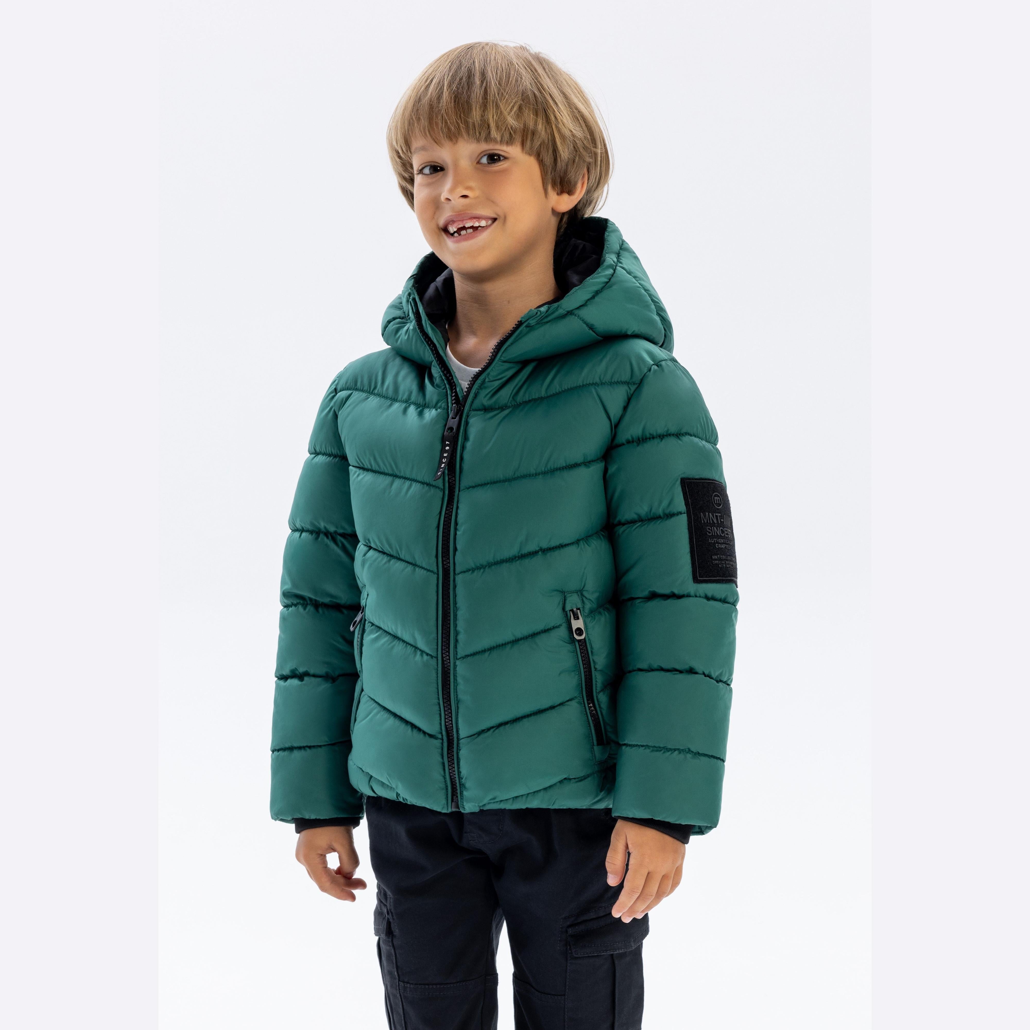 Young boy wearing green padded jacket with hood, standing against white background, smiling at camera
