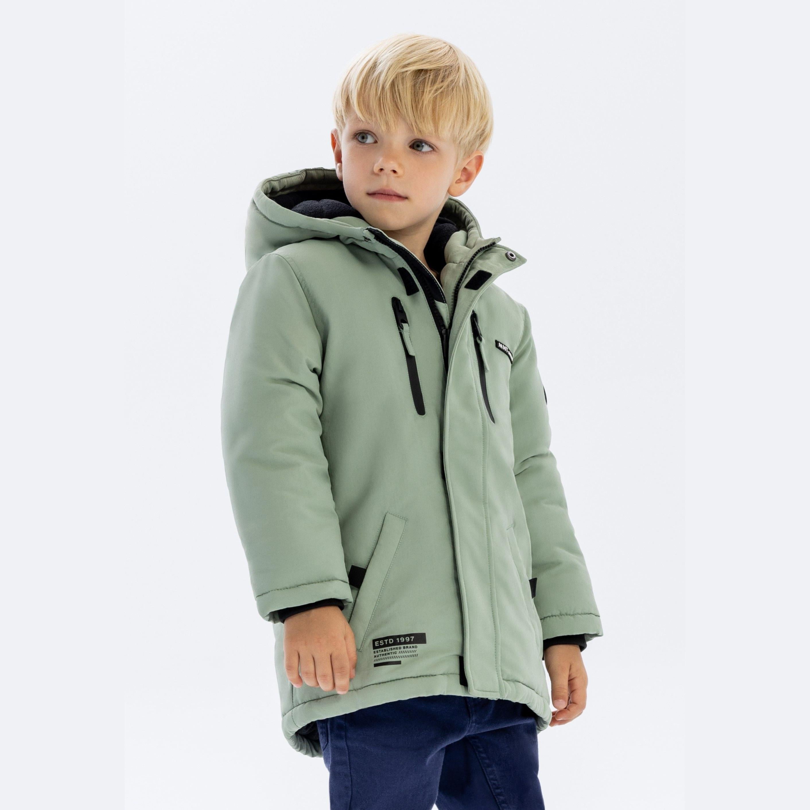 Young blonde boy wearing sage green parka jacket with hood up, hands in pockets, blue jeans, standing against white background