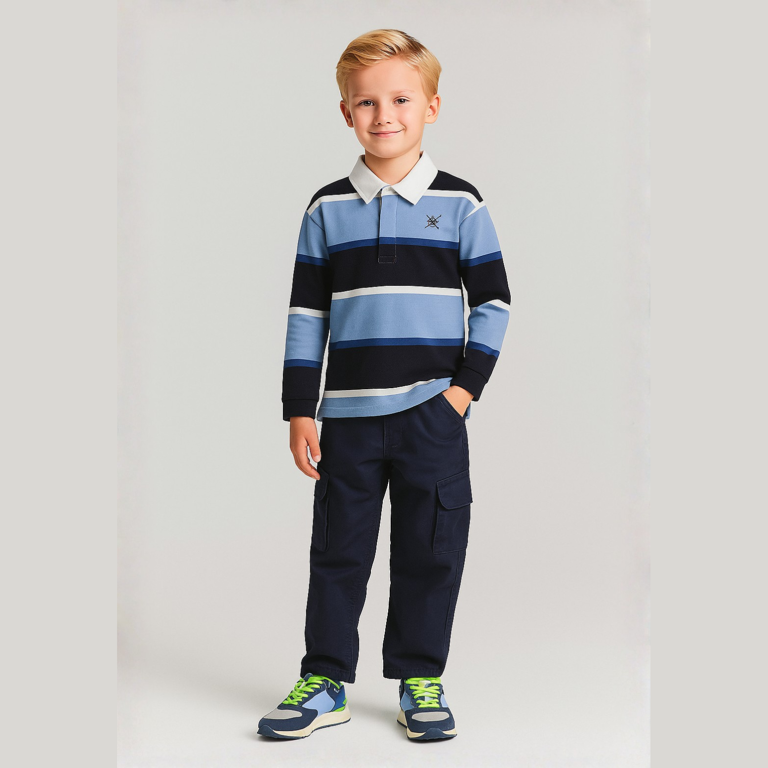 Young blonde boy wearing navy striped rugby polo shirt with white collar and dark trousers, standing against light background, full body front view
