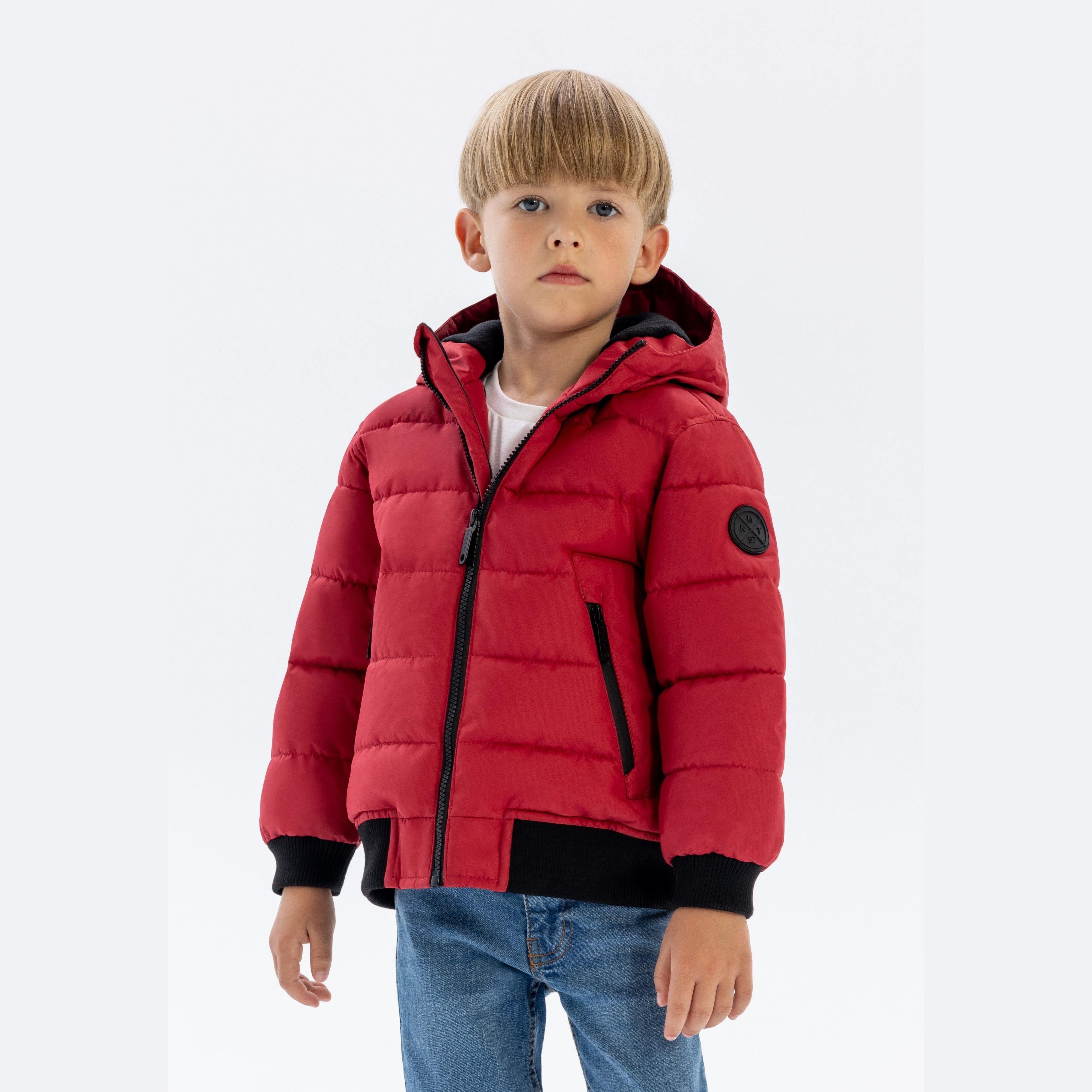 Young boy wearing red quilted padded jacket with black rib details, hood up, paired with blue jeans on white background