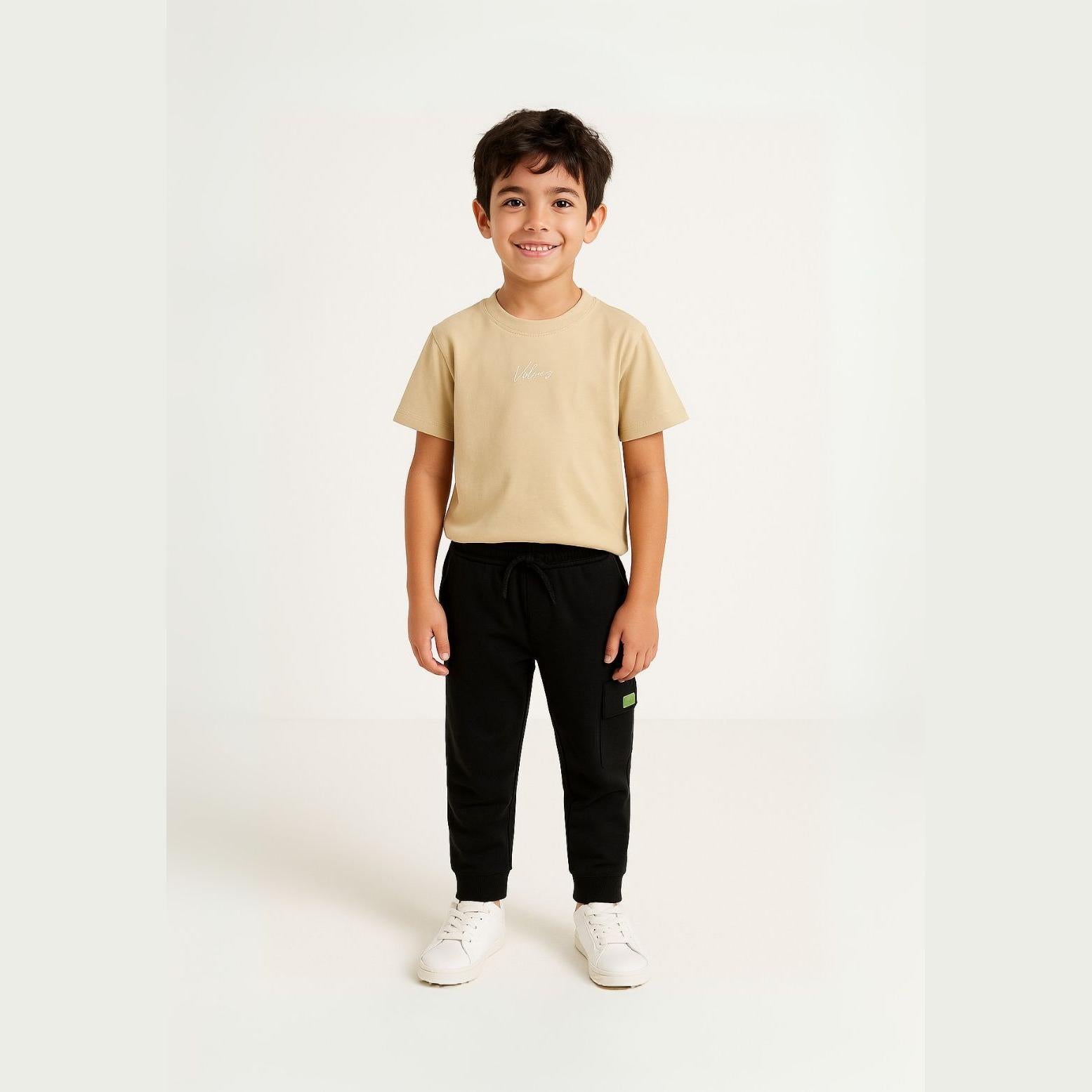 Young boy wearing sand coloured t-shirt with black joggers standing against white background