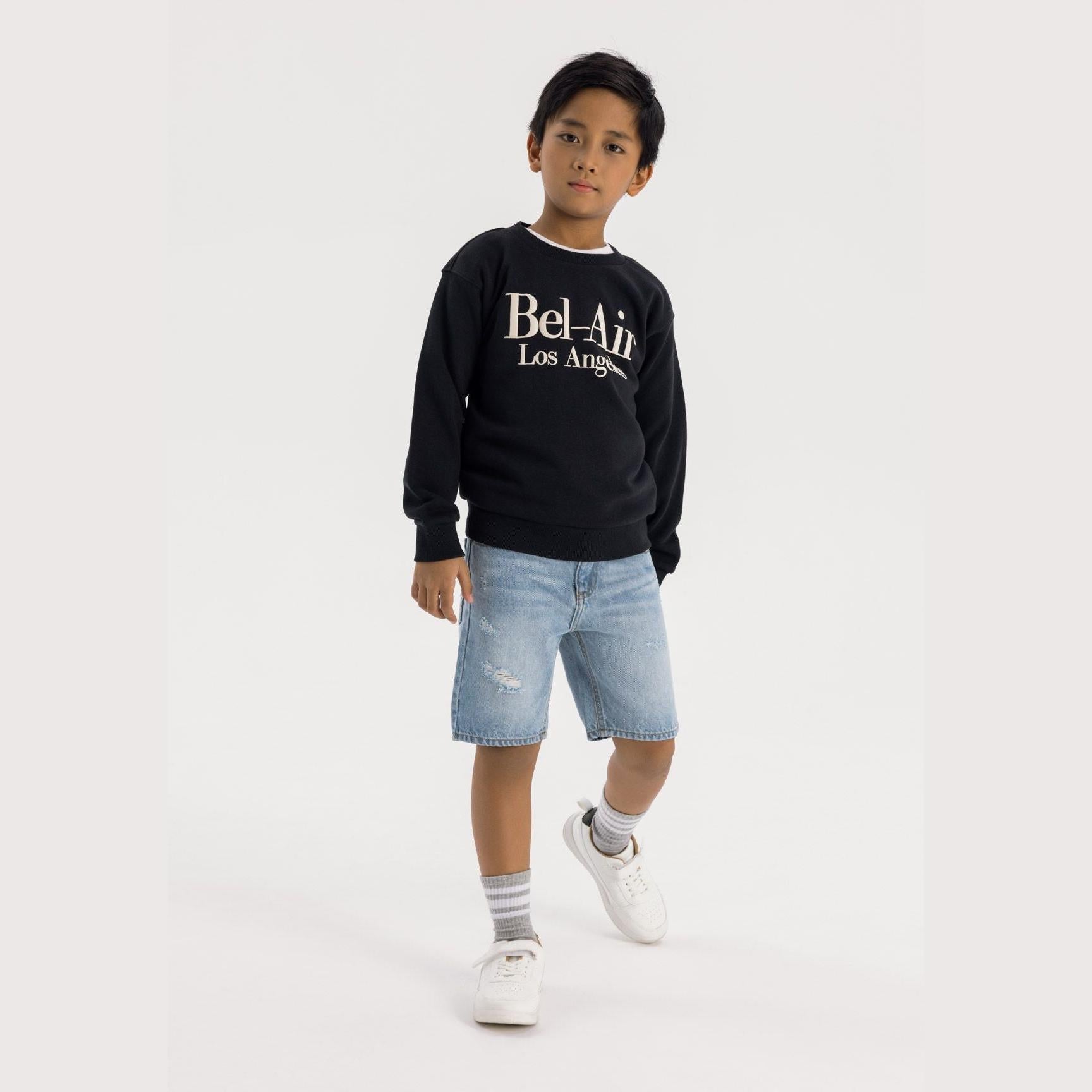 Young boy wearing black oversized crew neck sweatshirt with white Bel-Air Los Angeles print, paired with light blue denim shorts and white sneakers, standing against white background