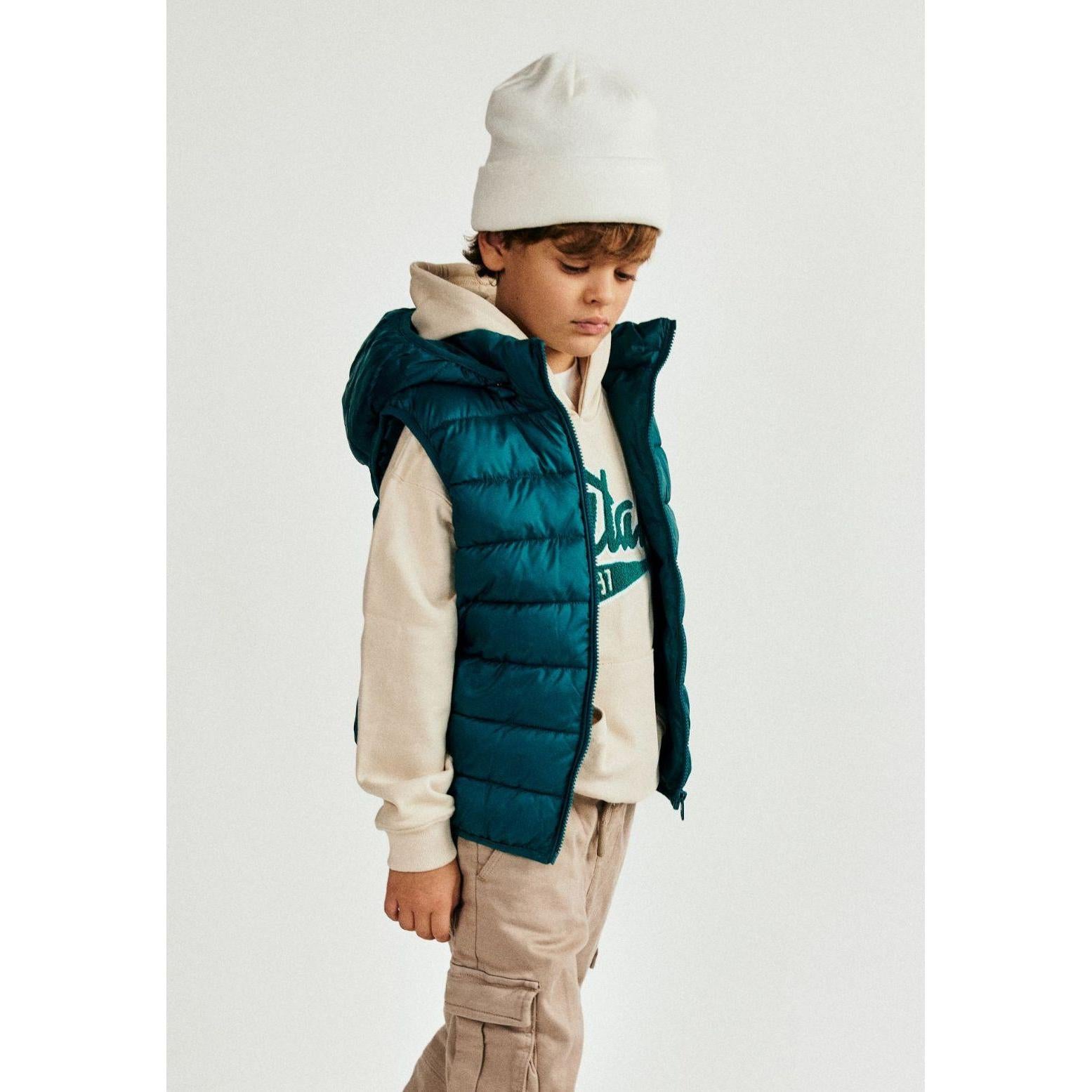 Young boy wearing green padded gilet with cream hoodie and beige cargo trousers, white beanie, standing against light grey background