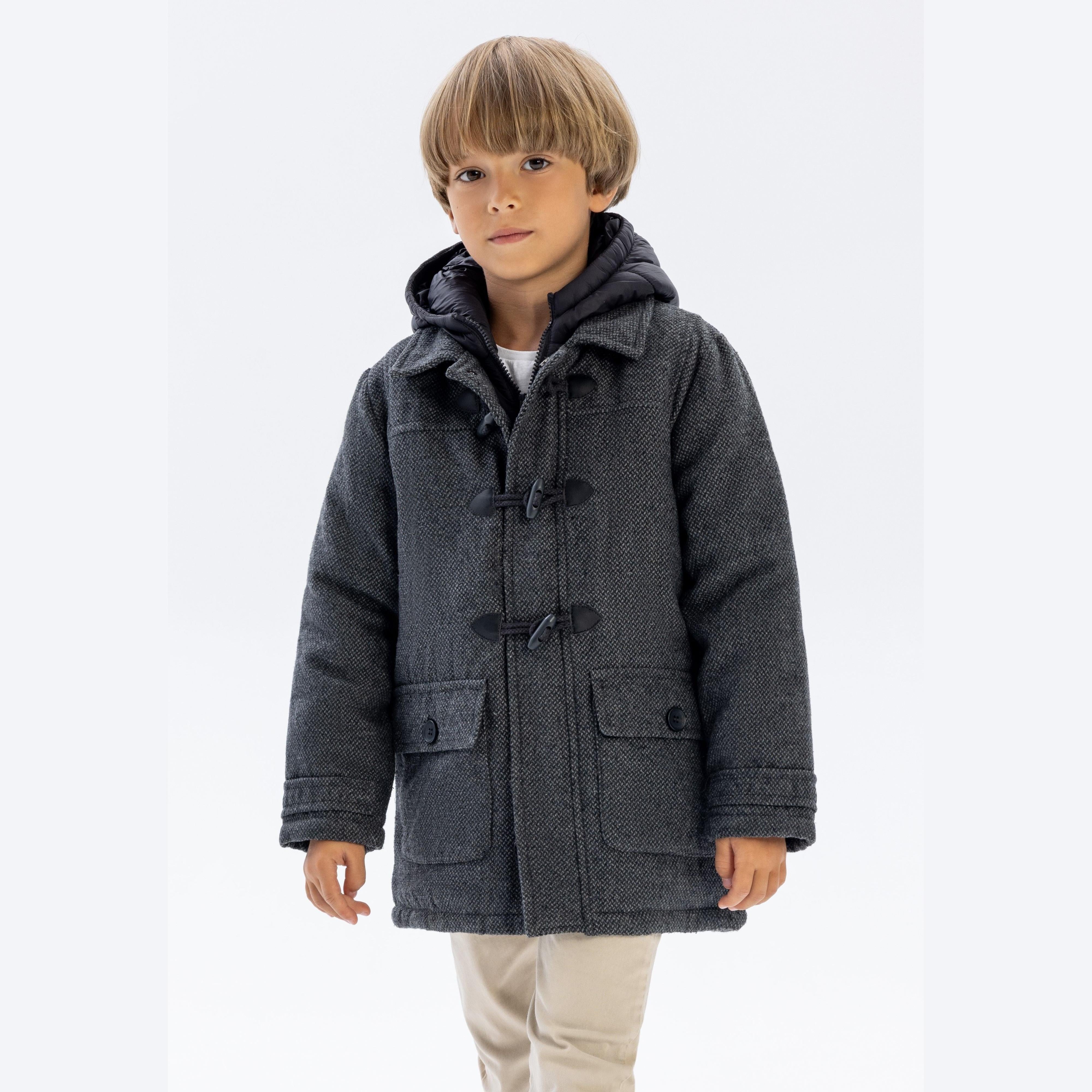 Young boy wearing grey tweed duffle coat with nylon hood and toggle fastenings, standing against white background