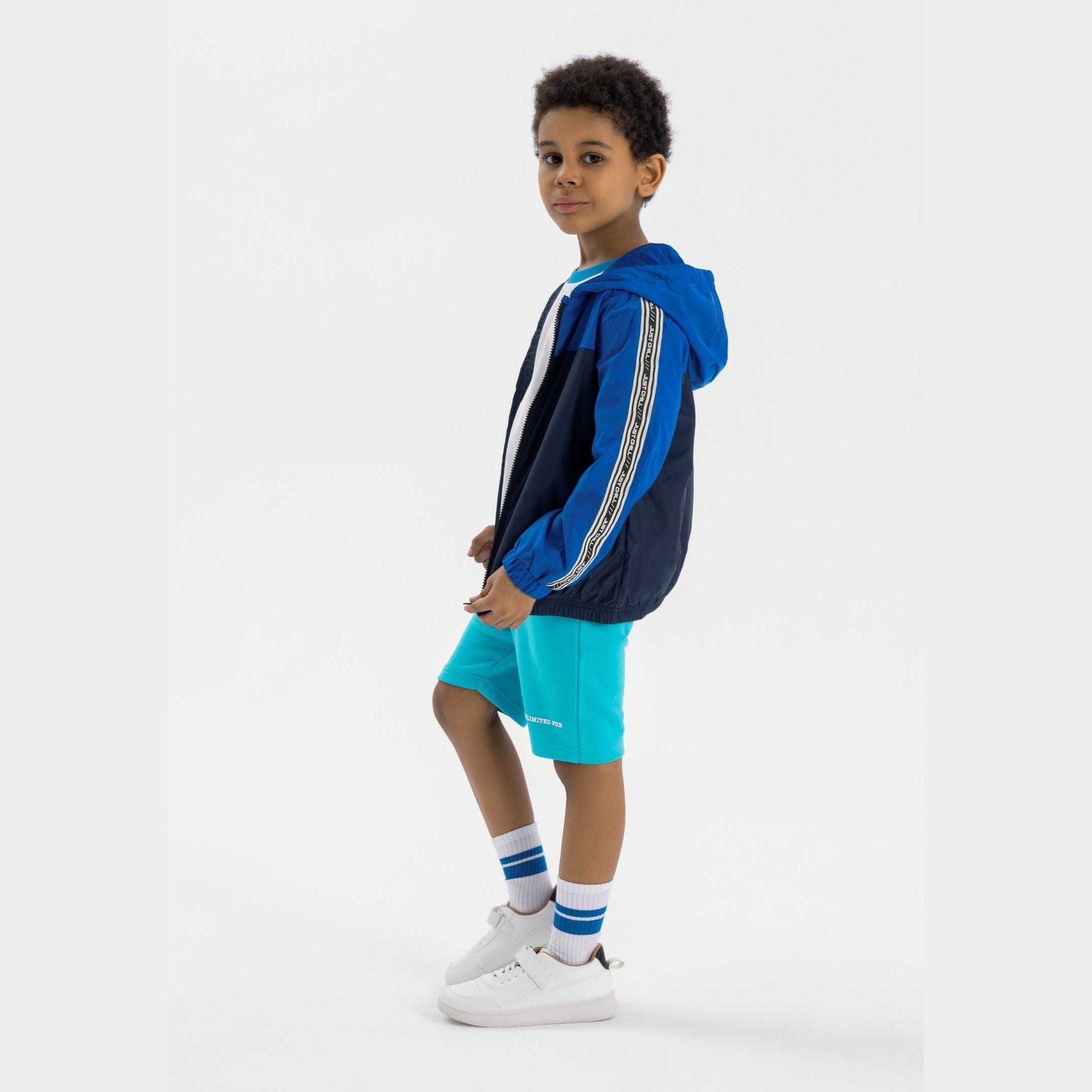 Young boy wearing cobalt blue and navy windbreaker jacket with hood, paired with turquoise shorts and striped socks, standing against white background