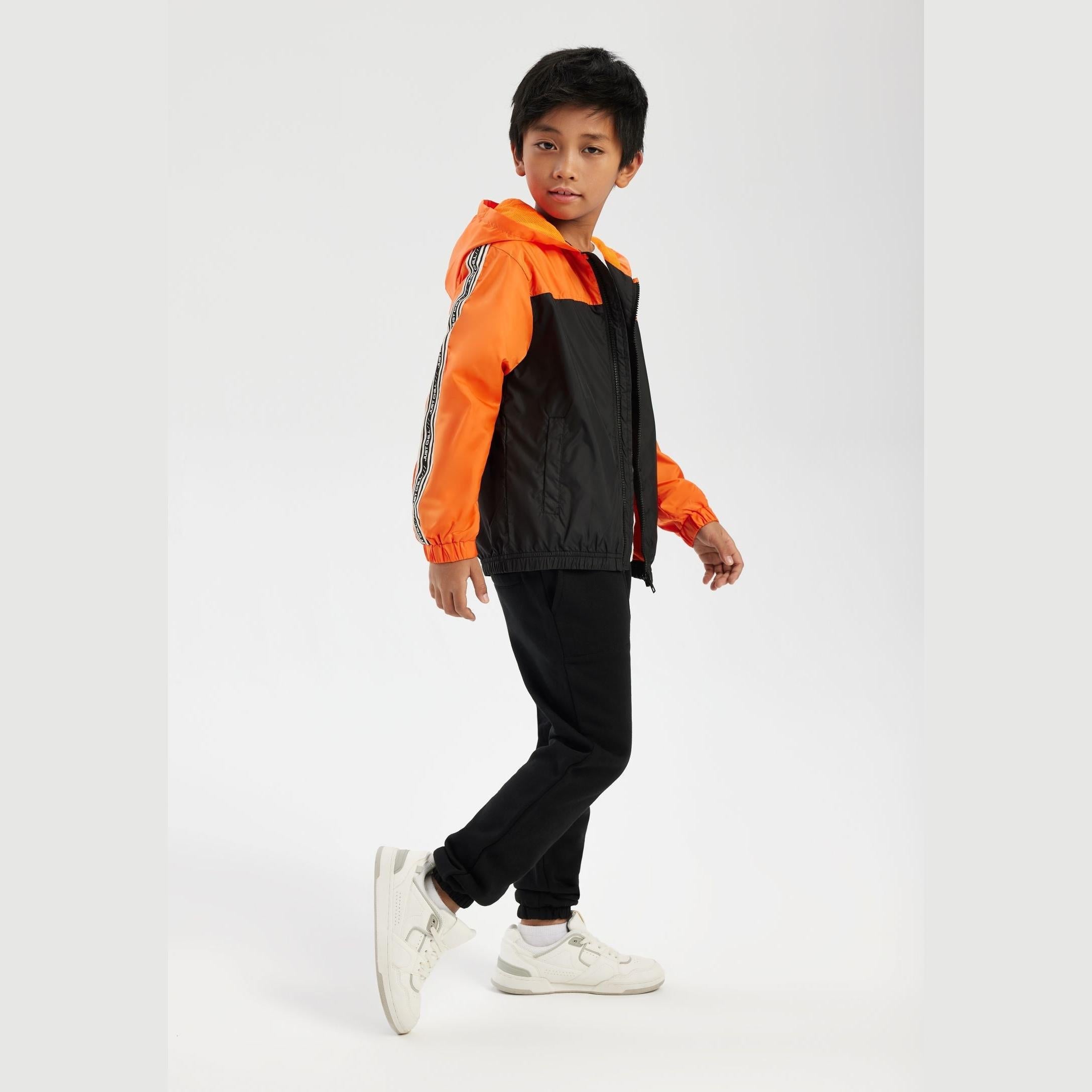 Young boy wearing bright orange and black colorblock windbreaker jacket with hood, paired with black joggers and white sneakers, standing against white studio background