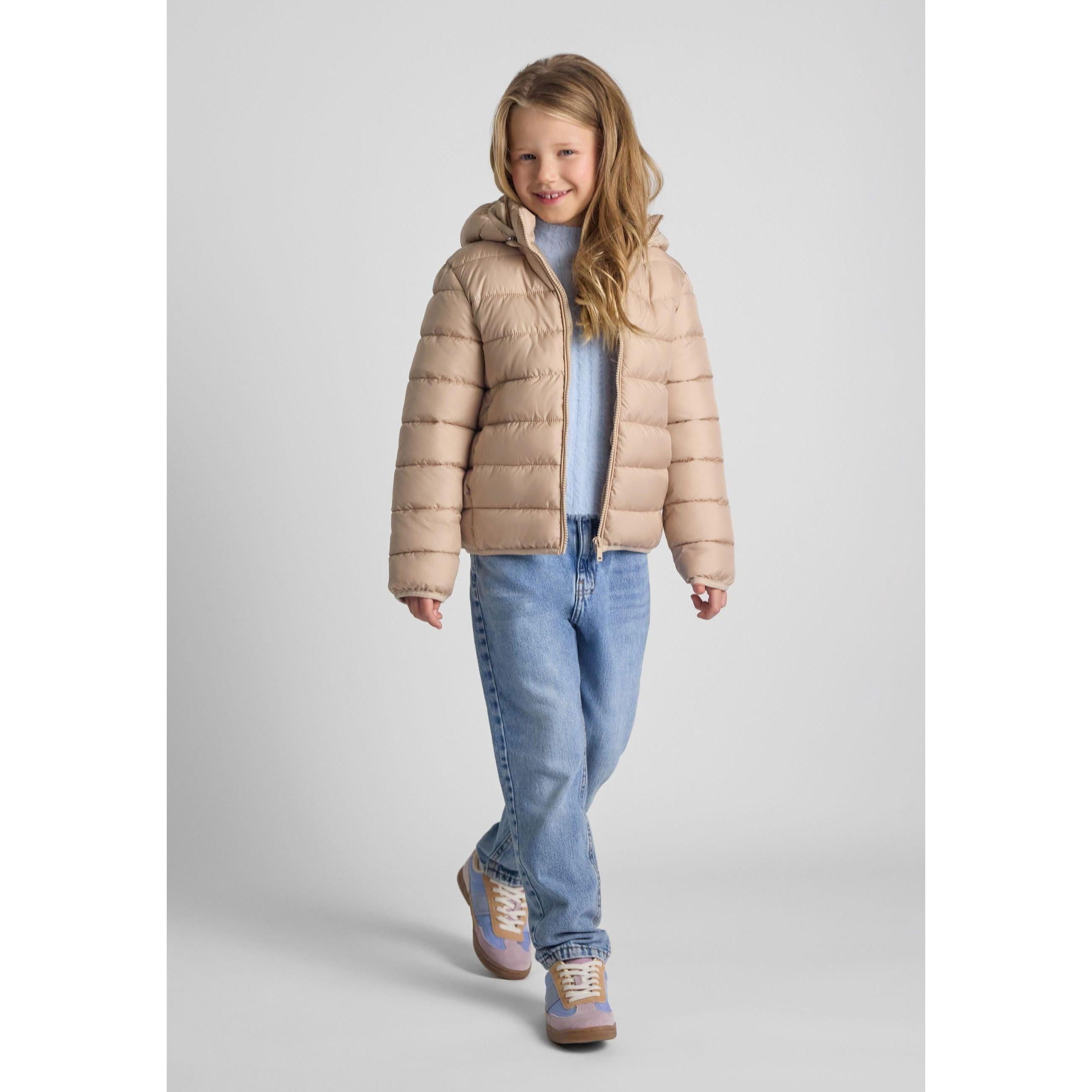 Young blonde girl wearing beige quilted jacket with detachable hood, light blue top and jeans, walking pose against white studio background