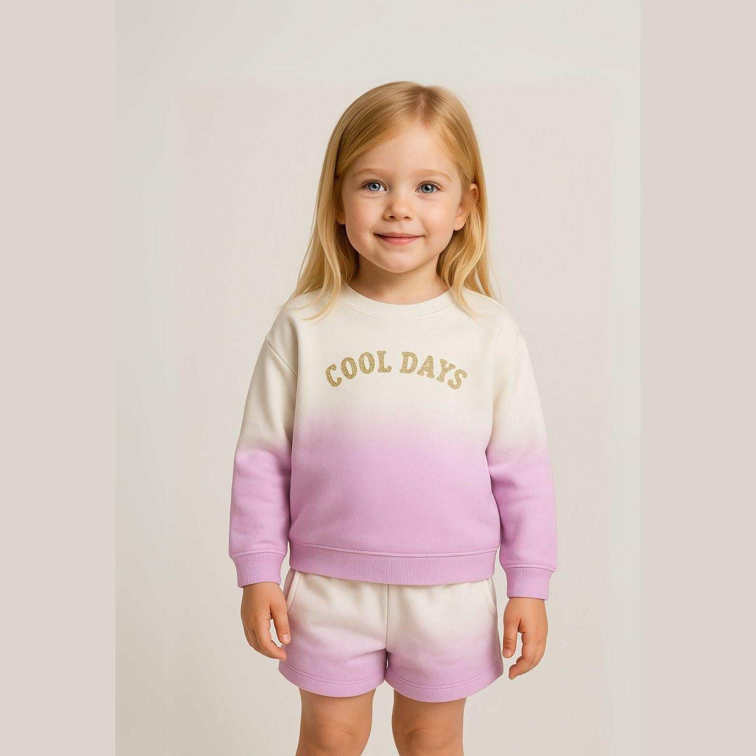 Young blonde girl wearing purple ombré fleece sweatshirt and shorts set with 'Cool Days' glitter text, standing against neutral background