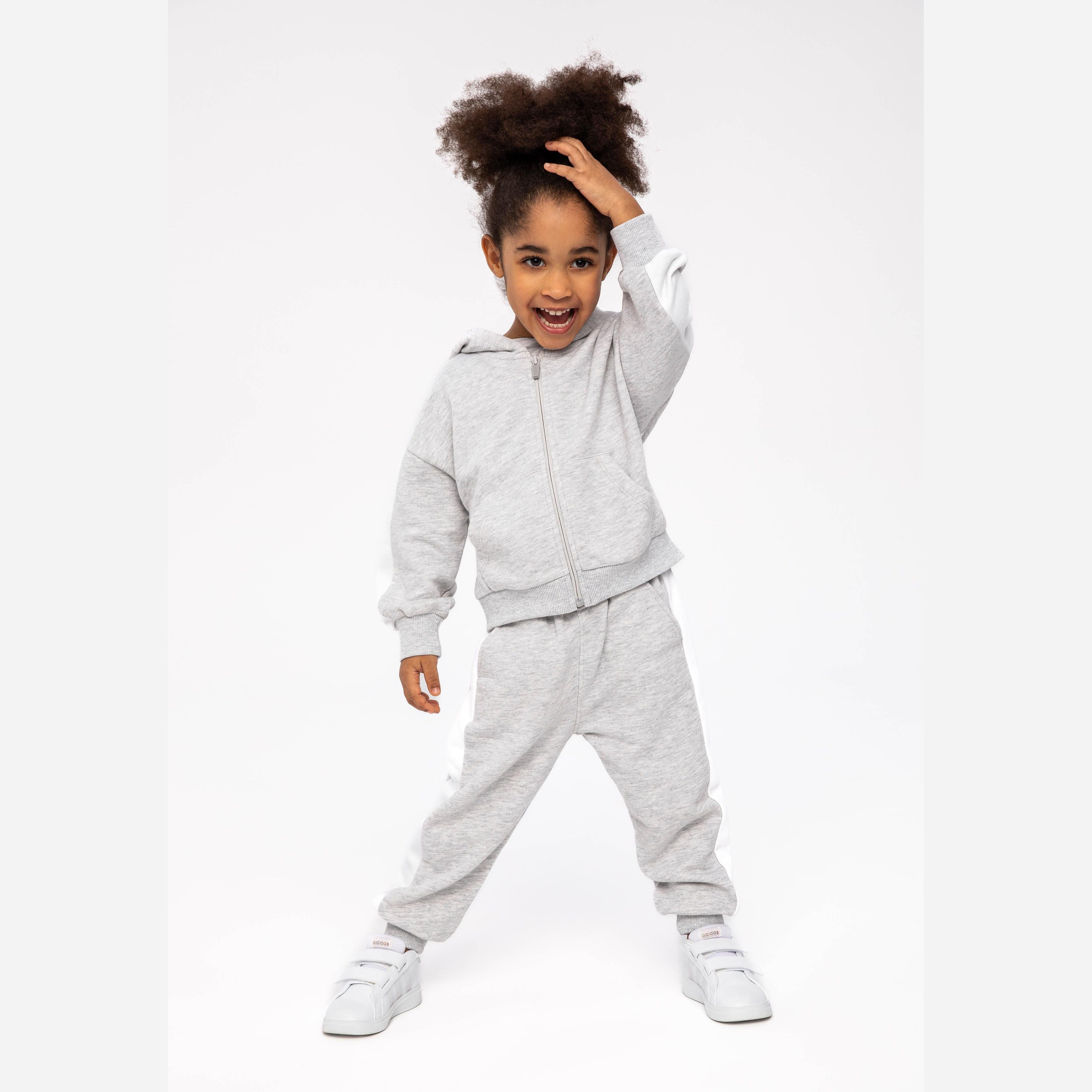 Young girl wearing grey zip-up hoodie and matching joggers, posing playfully with hand on head against white studio background