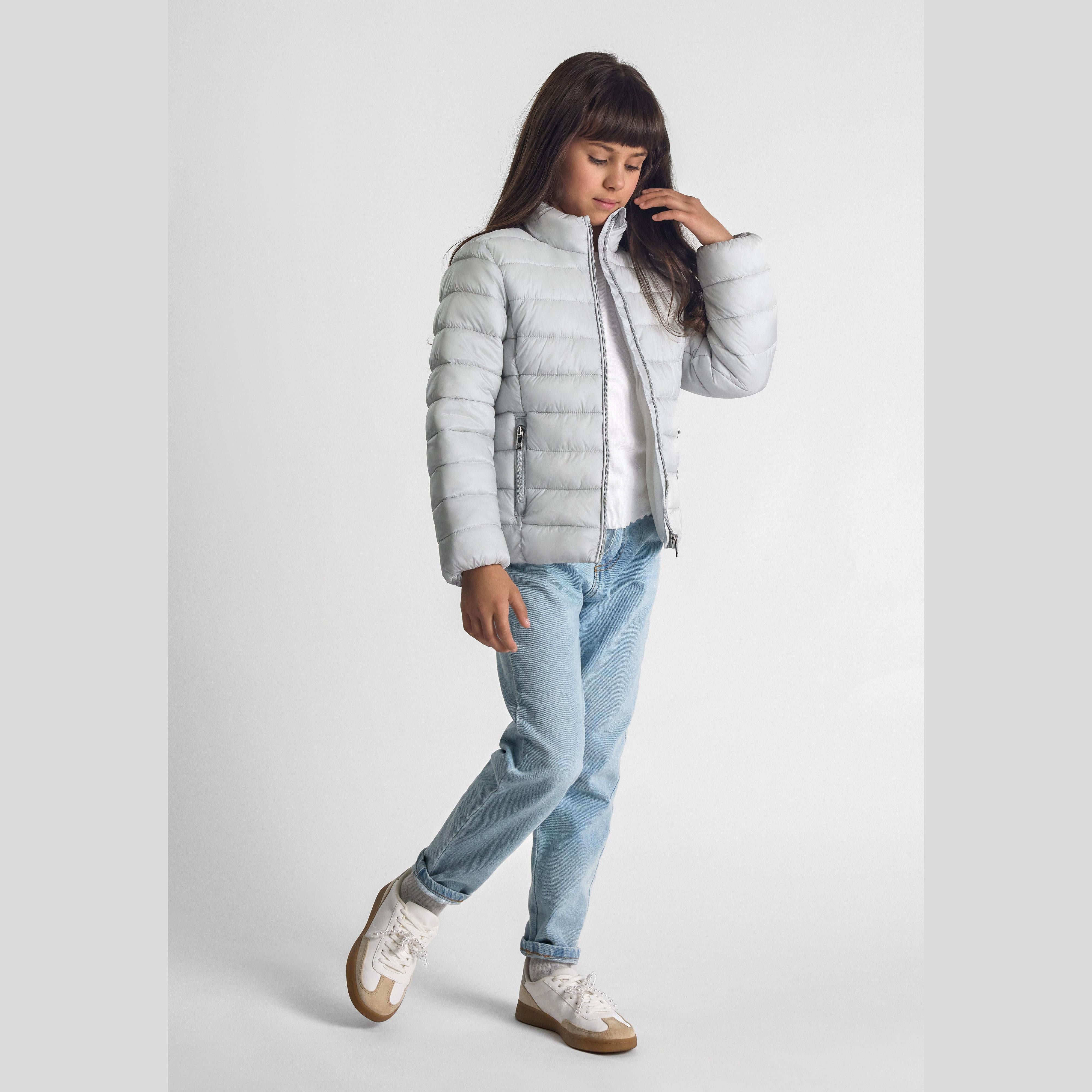 Girl wearing silver-grey quilted jacket with blue jeans and white sneakers on white background, showing full outfit styling