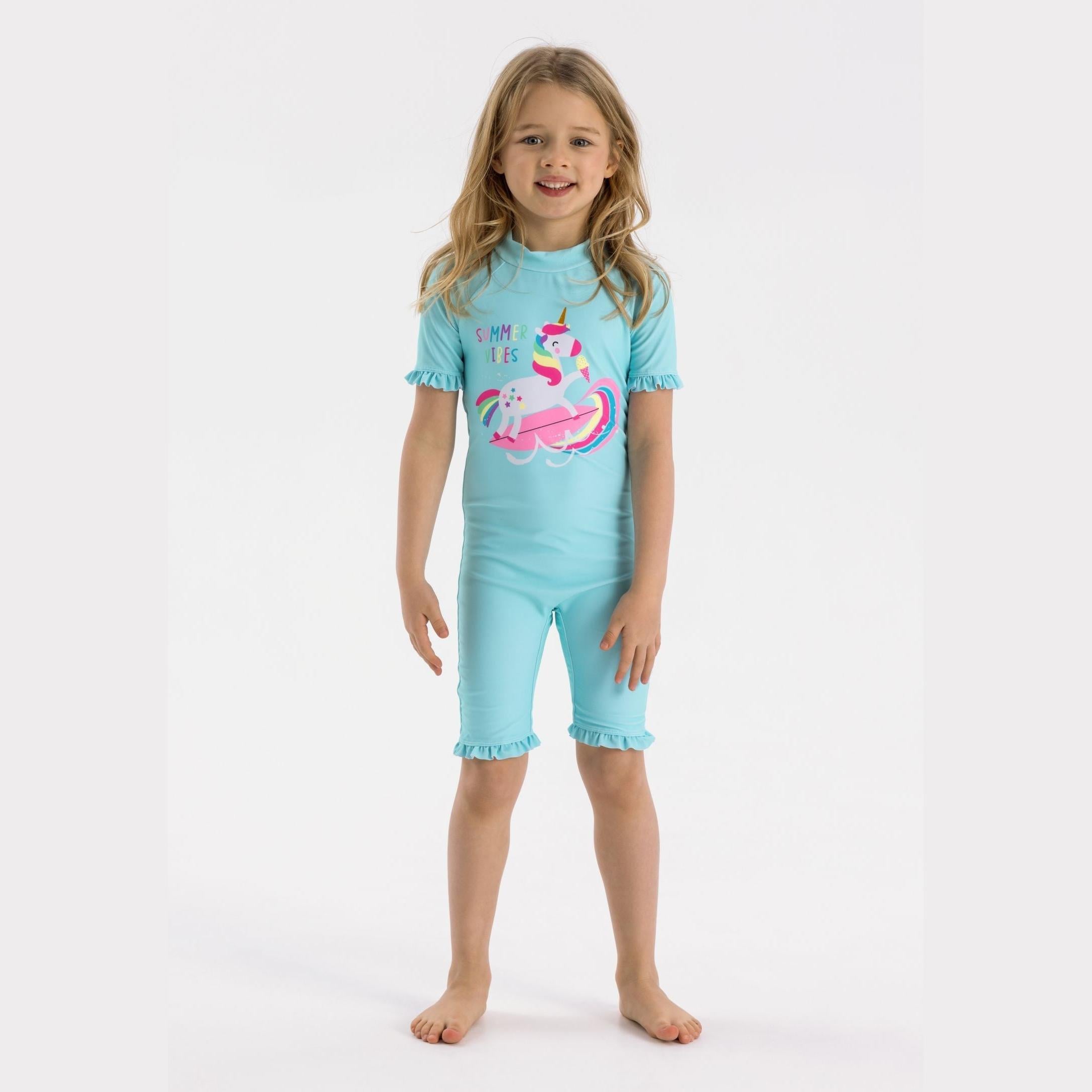 Young girl wearing light blue rash suit with unicorn print and matching ruffle details, standing against white background