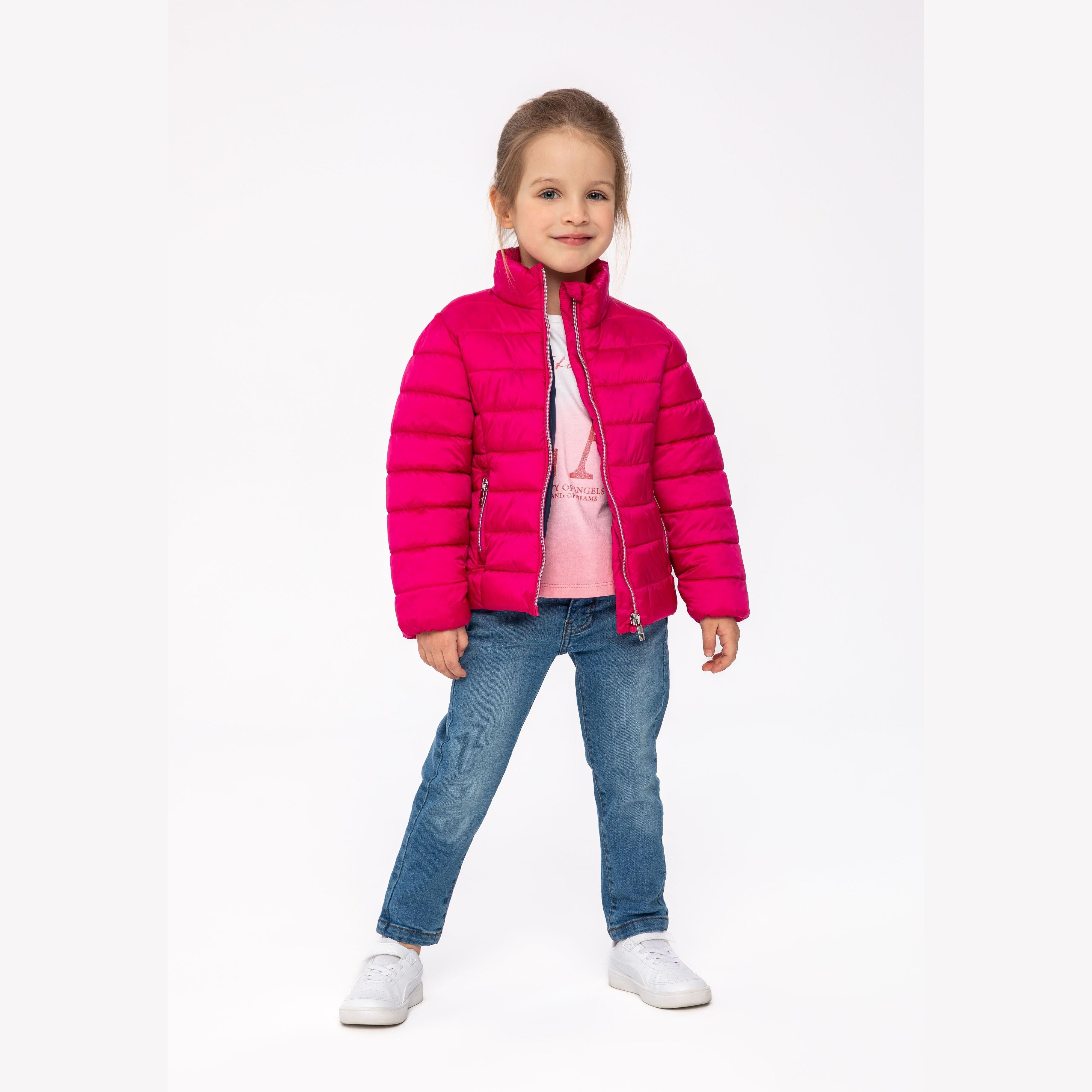 Young girl wearing bright pink quilted padded jacket over pink top and blue jeans, standing against white background, smiling at camera