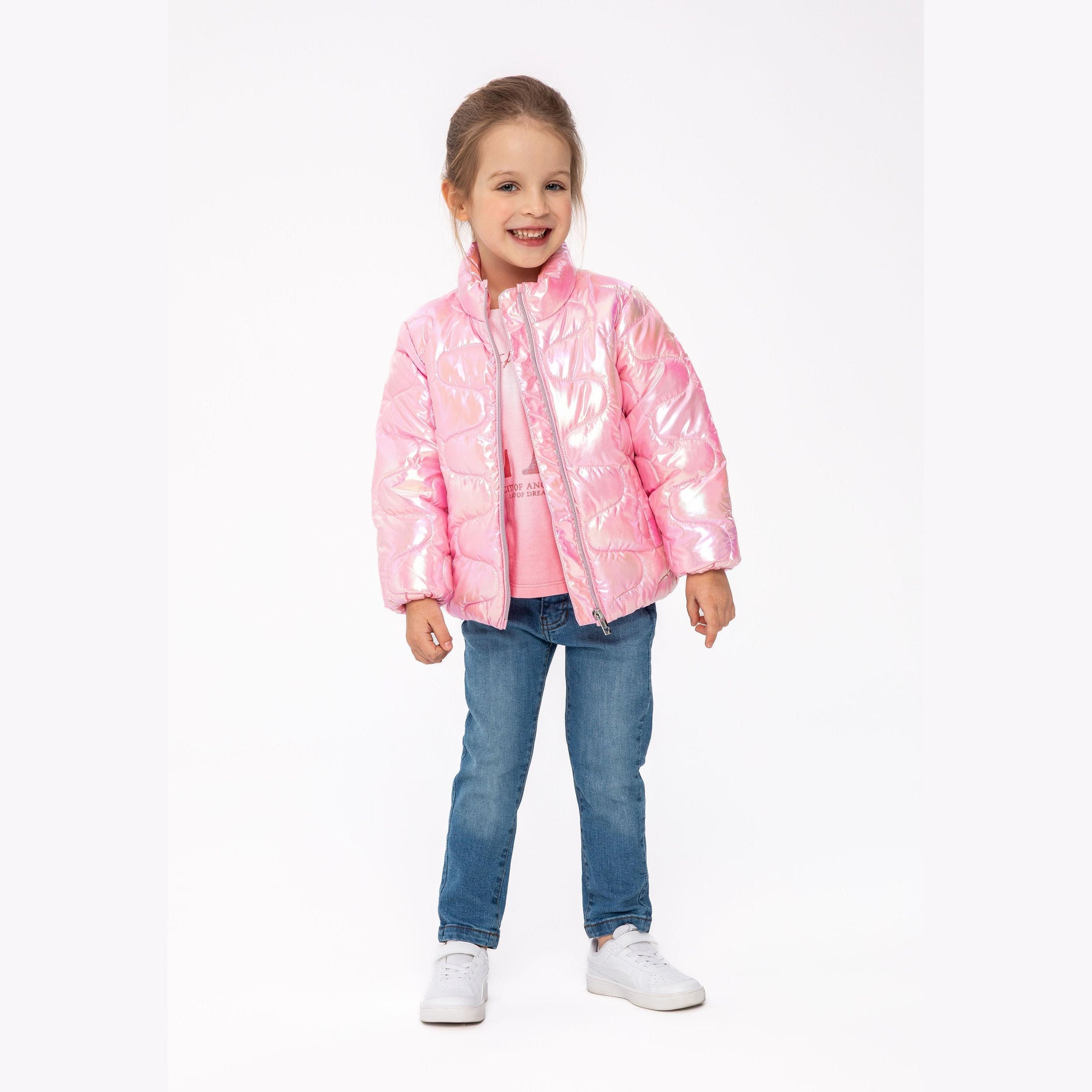 Young girl wearing shimmery pink padded jacket with funnel neck, paired with blue jeans and white sneakers on light background