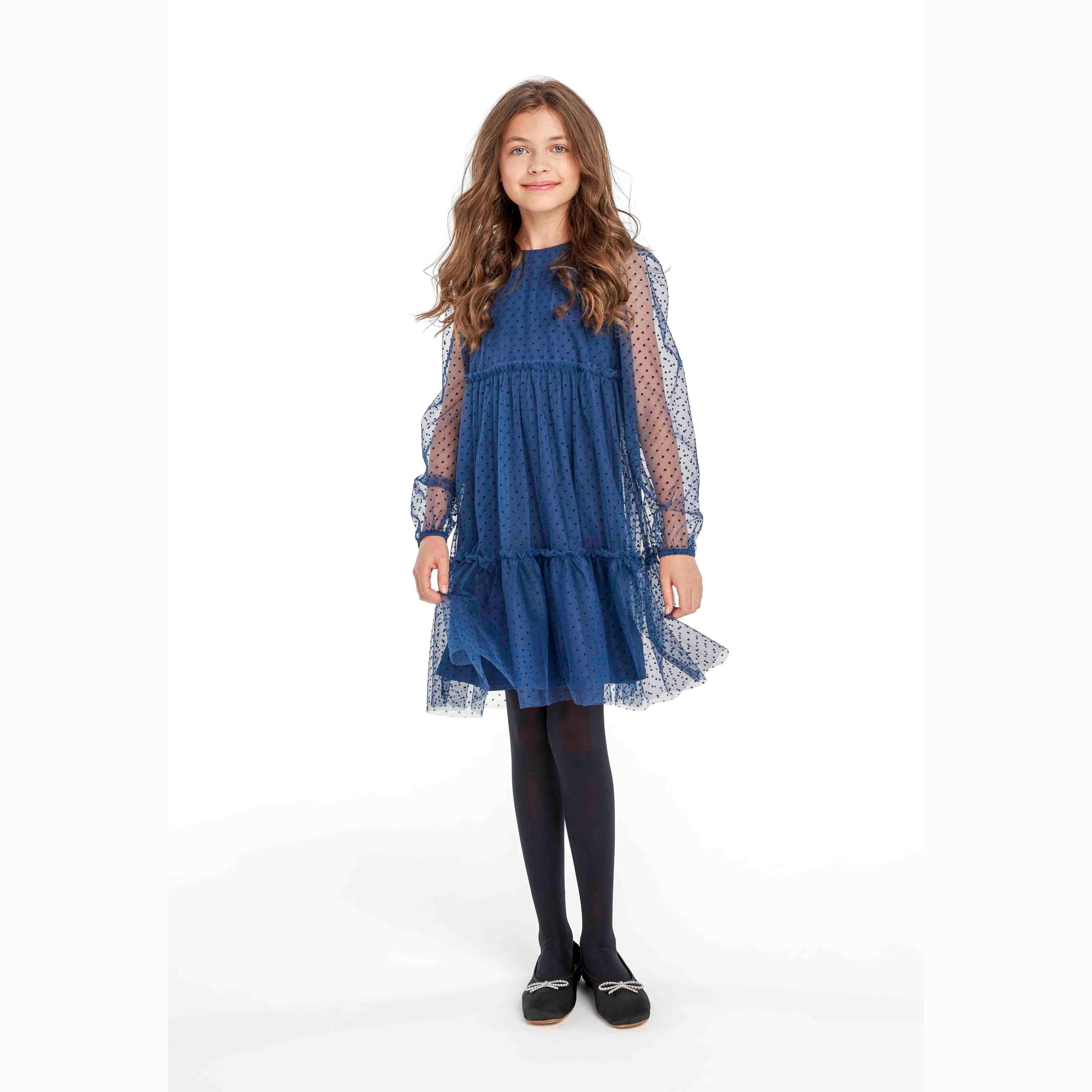 Young girl wearing navy blue party dress with flock print pattern, long balloon sleeves and tiered skirt, paired with black tights and shoes, standing against white background