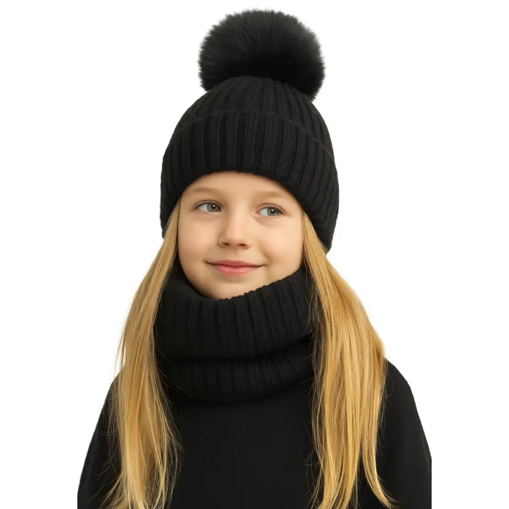Young blonde girl wearing black ribbed knit hat with faux fur pom pom and matching black knitted snood, smiling against white background