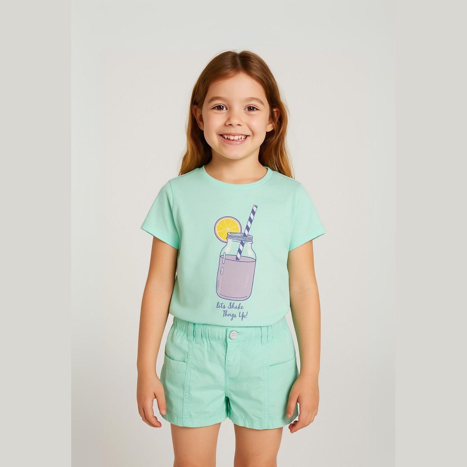 Young girl wearing mint green t-shirt with smoothie print and matching mint poplin shorts, standing against white background