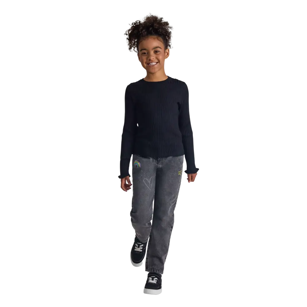 Young girl wearing black ribbed jumper with frill cuffs and grey jeans, full body shot on black background