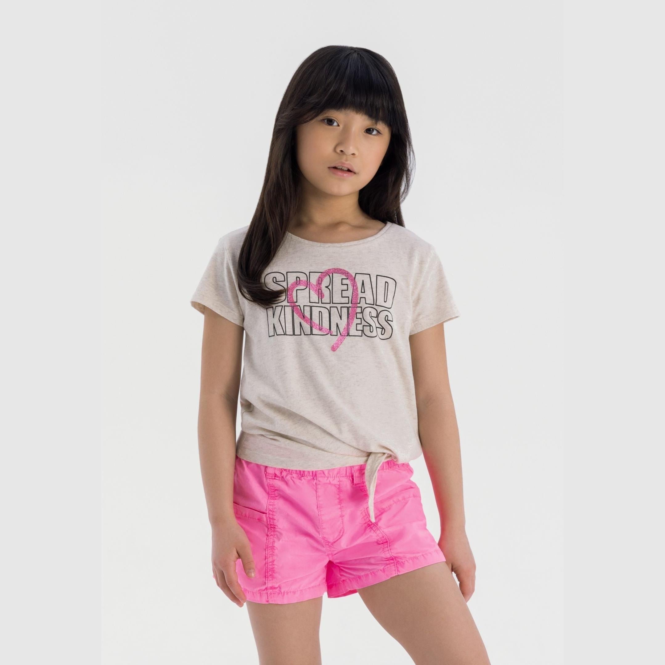 Young girl wearing beige tie-front t-shirt with Spread Kindness text and pink heart, paired with bright pink shorts, standing against white background