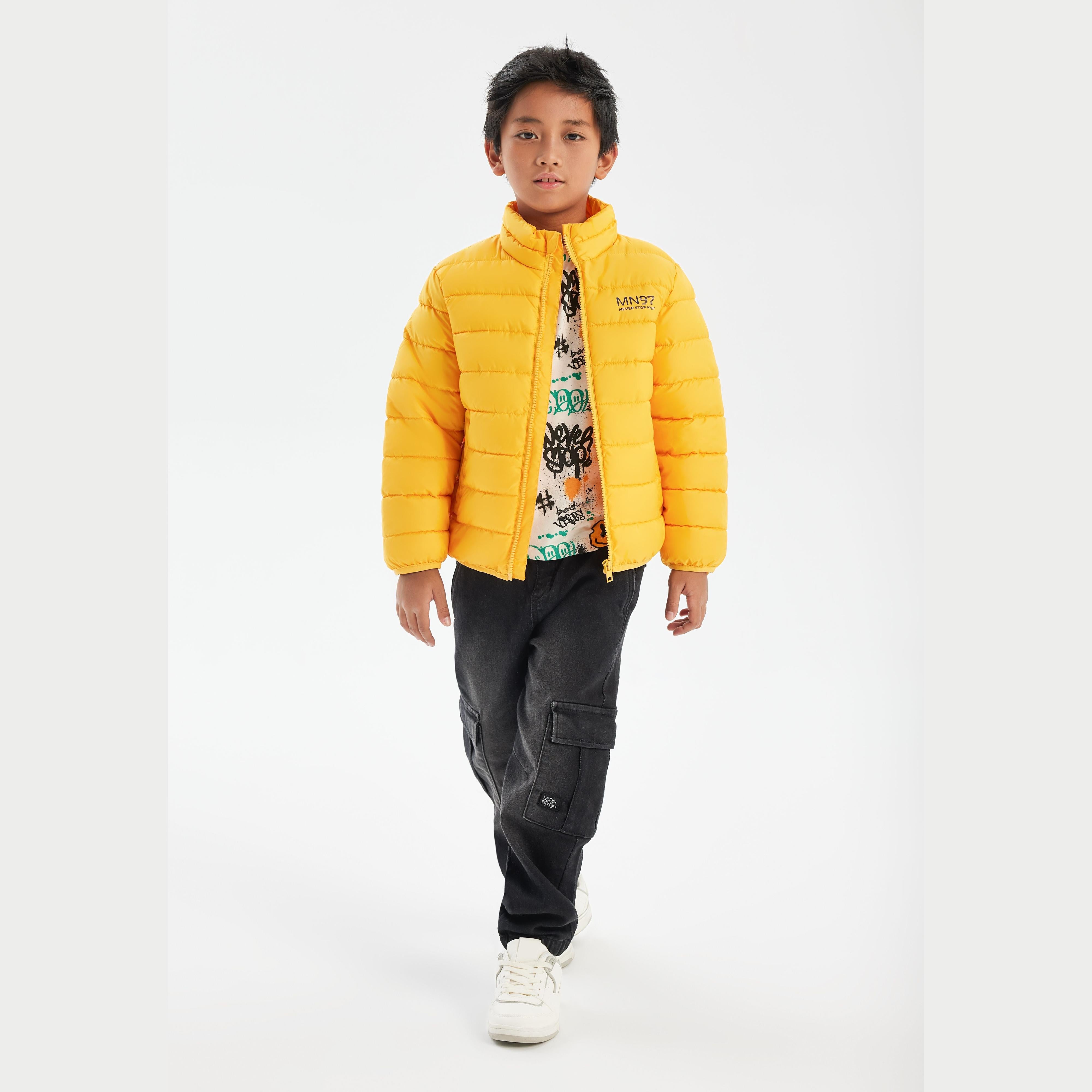 Boy wearing bright yellow quilted jacket with zip closure, standing against white background, paired with graphic t-shirt and dark cargo pants