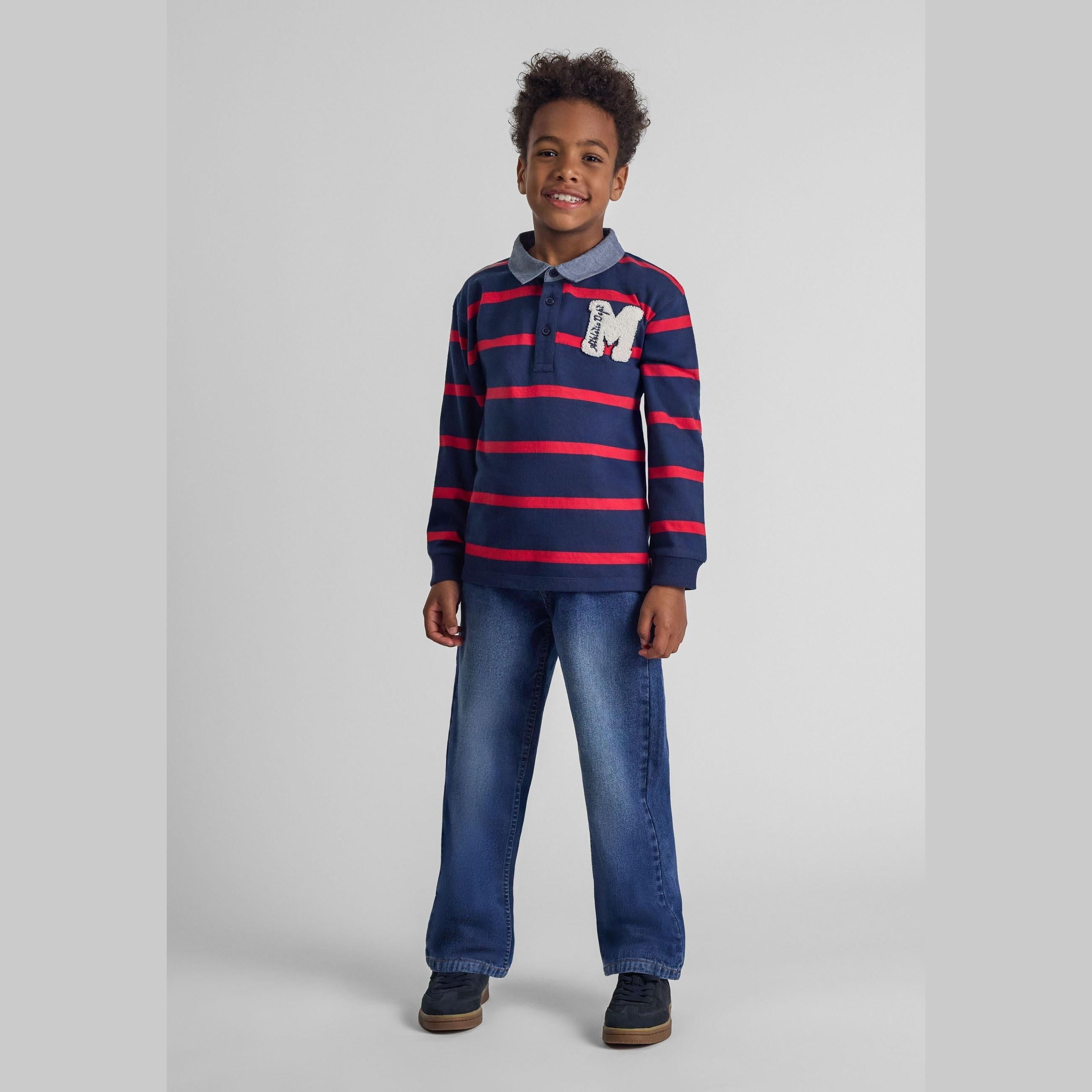 Young boy wearing navy striped long sleeve rugby shirt and baggy fit navy jeans, standing against grey background