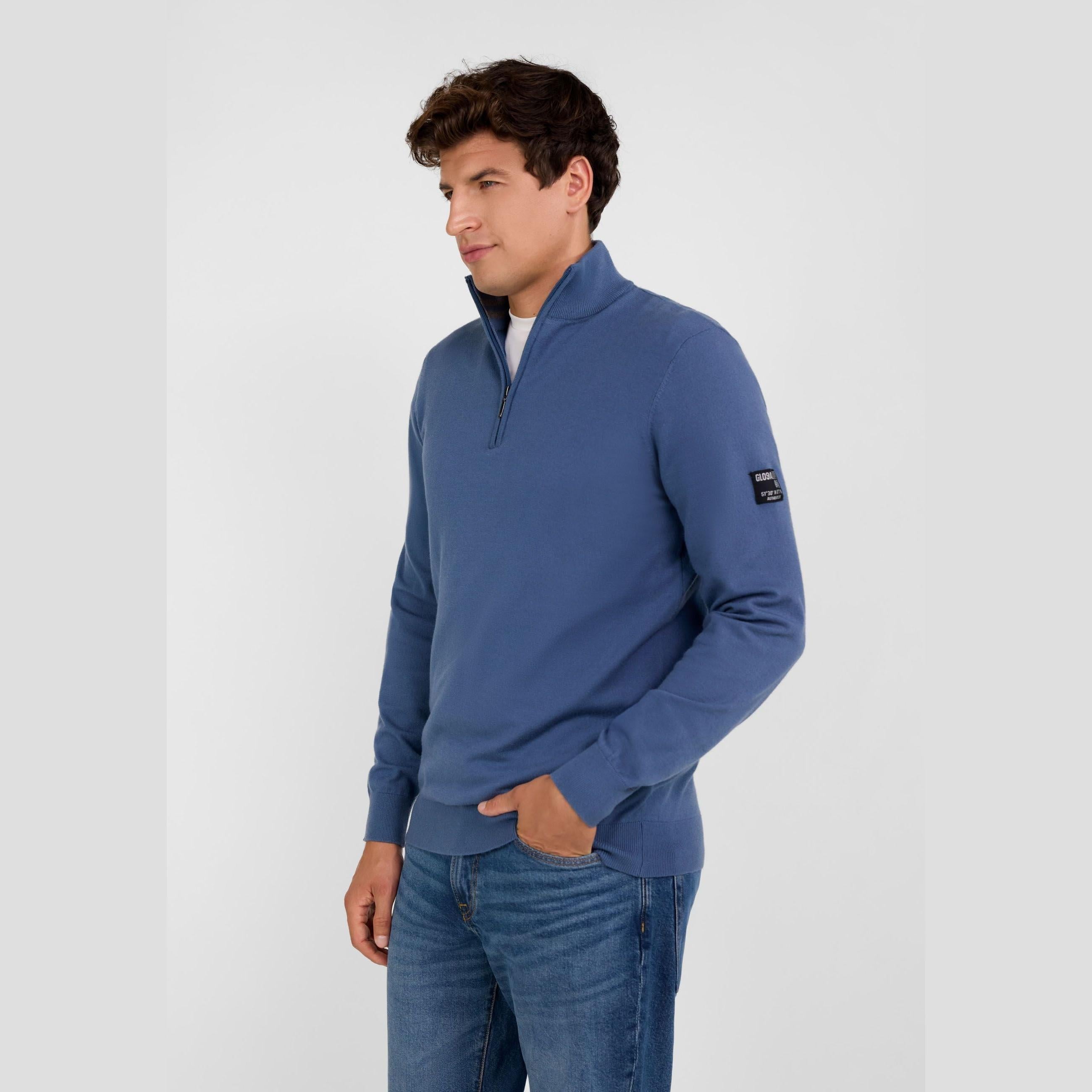 Man in blue half zip jumper shown from side angle, relaxed pose with hands in pockets, white studio background