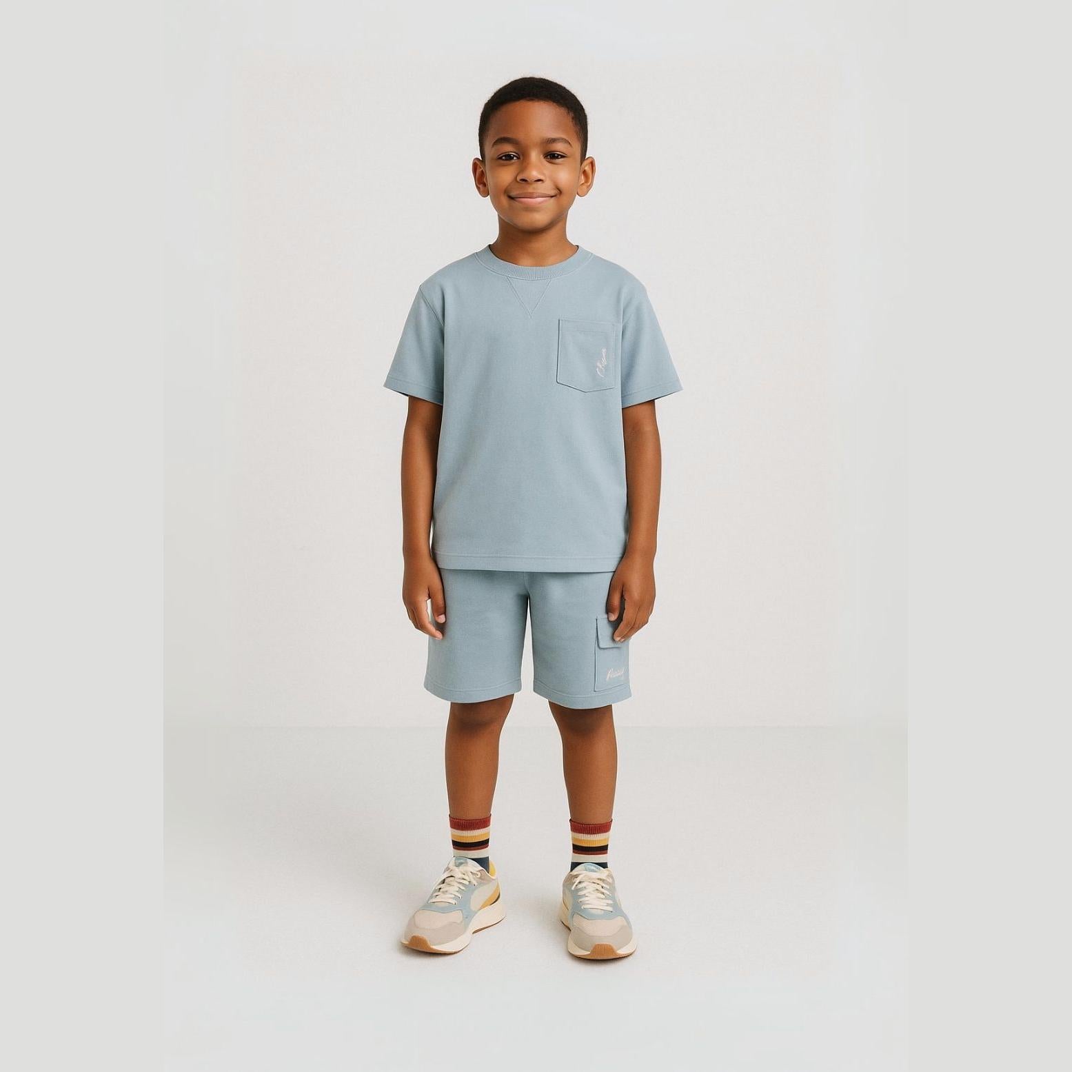 Young boy wearing light blue t-shirt and shorts set standing against white background, complete with striped socks and trainers