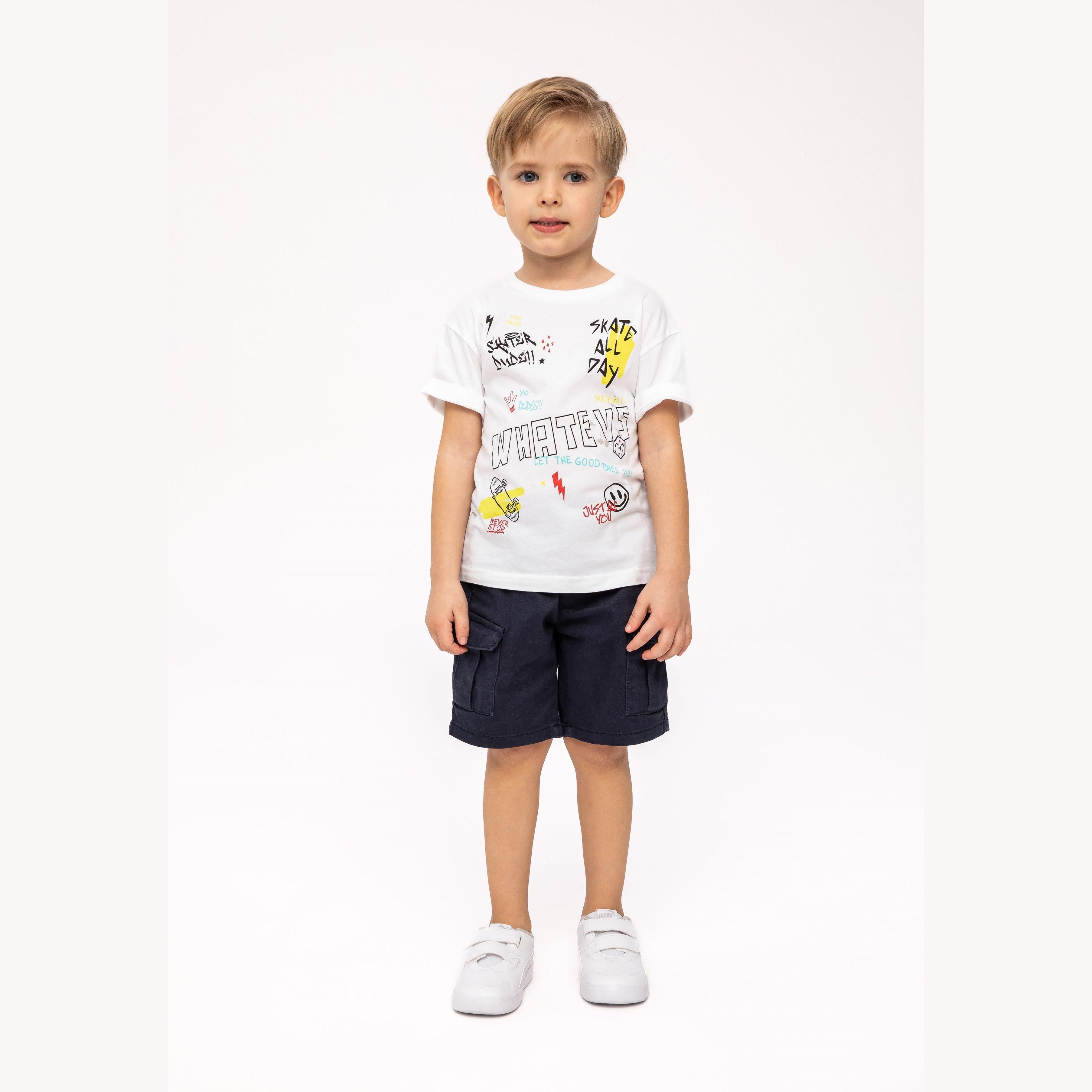 Young boy wearing white graphic t-shirt with skateboard print and navy shorts, standing against light background