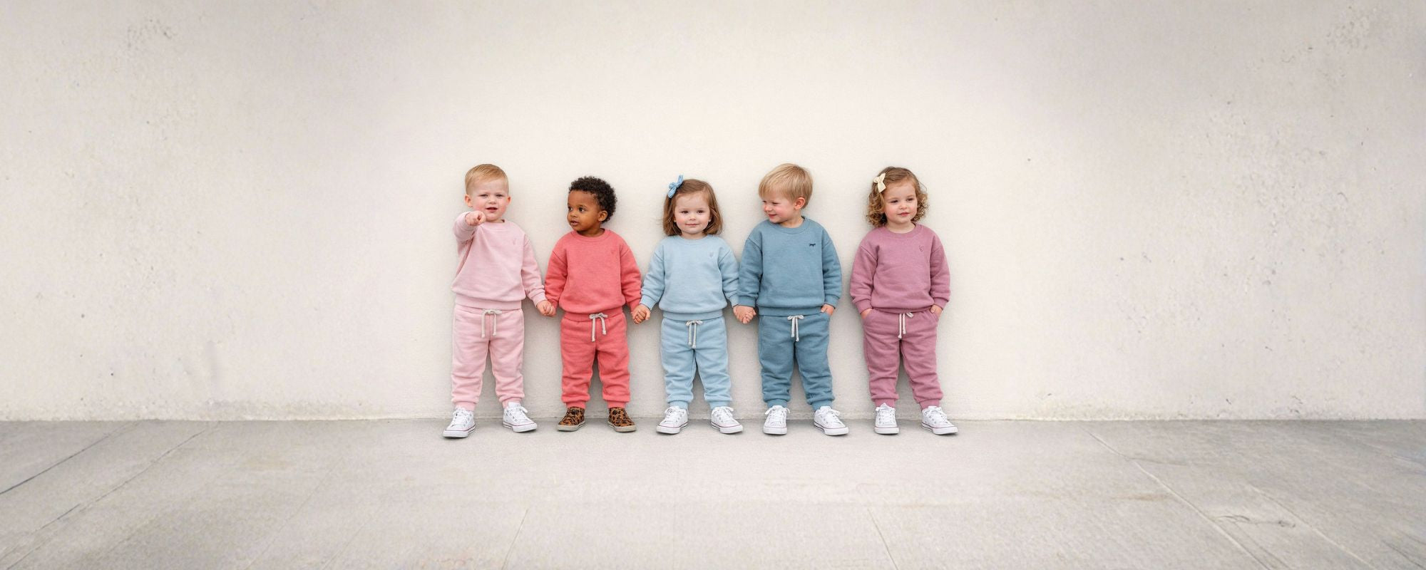 Six toddlers standing in a row against a neutral wall, wearing matching pastel-colored tracksuits in pink, coral, light blue, and mauve, holding hands and facing forward.