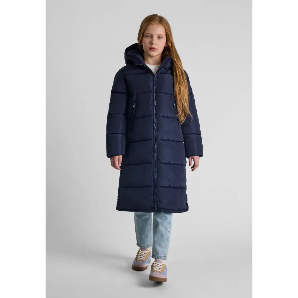 Girl wearing navy quilted long winter coat with hood, paired with light blue jeans and brown sneakers against white studio background