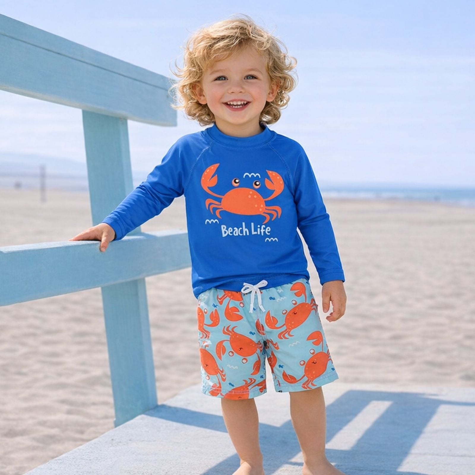 Smiling toddler in a blue crab-print swim set stands by a beach railing, capturing a fun, playful seaside vibe.
