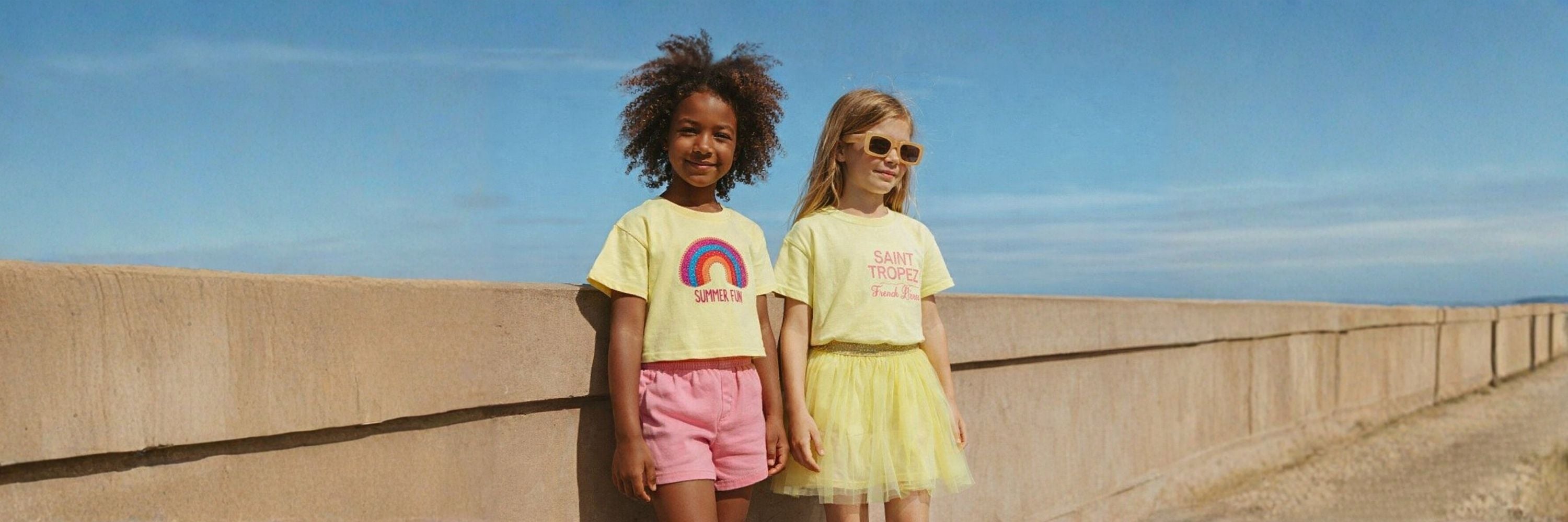 Two girls stand by a seaside wall in bright summer outfits, enjoying a sunny day with a relaxed, cheerful vibe.