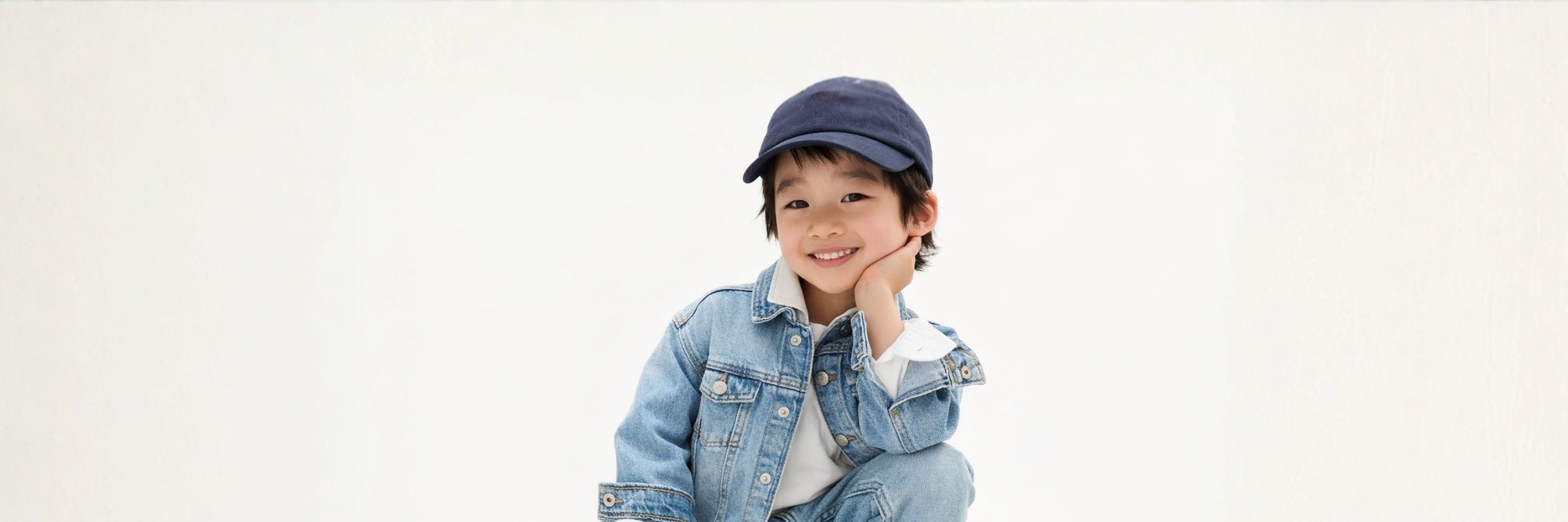 A smiling boy in a denim jacket and cap poses playfully with his hand on his cheek against a clean, minimal background.