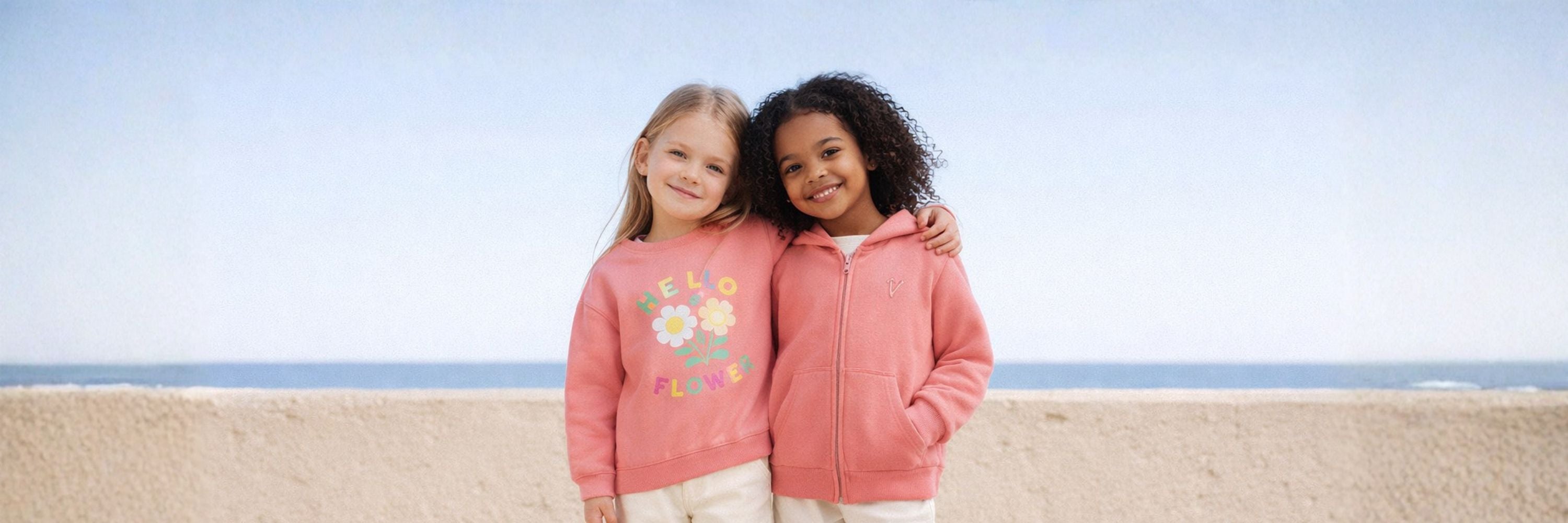 Two girls smiling by the beach with arms around each other, wearing pink sweatshirts—one with a flower print and the other a zip hoodie, with the sea in the background.