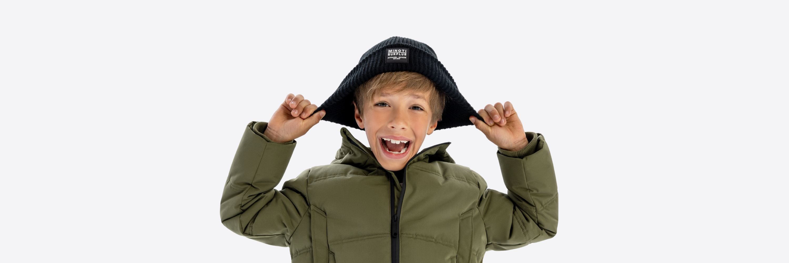A laughing teenage boy wearing a green puffer jacket, holding up the edges of a black beanie hat on his head against a white background.