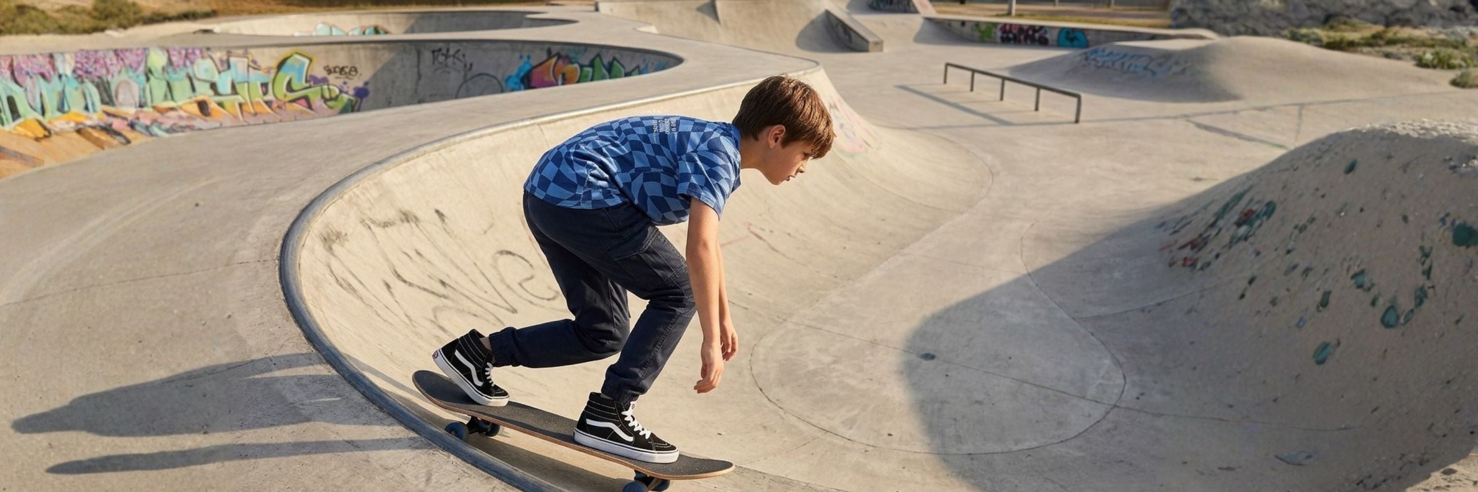 A boy skateboarding in a concrete skate park, wearing a blue checkered t-shirt, dark trousers, and high-top sneakers.
