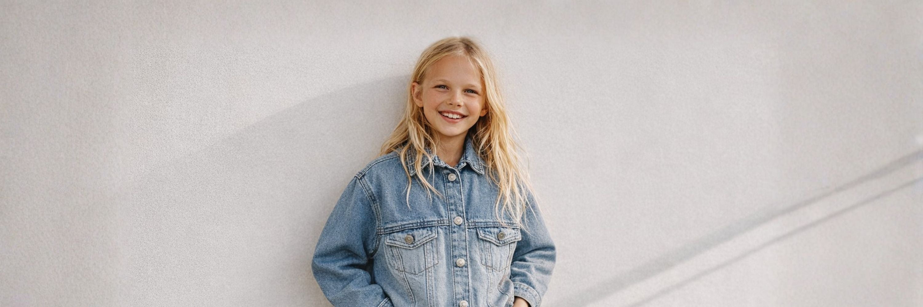 A smiling girl wearing a denim jacket stands against a light, textured wall, giving a relaxed and casual look.