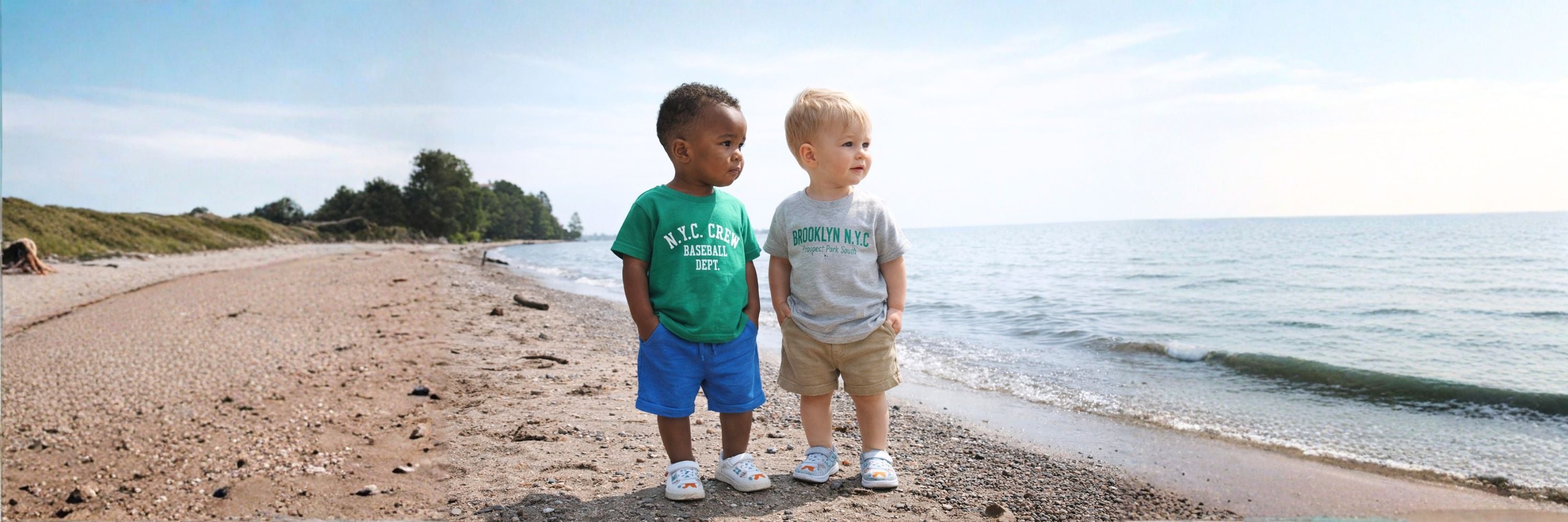 Two toddler boys stand side by side on a beach, looking into the distance in casual summer outfits as waves gently reach the shore.