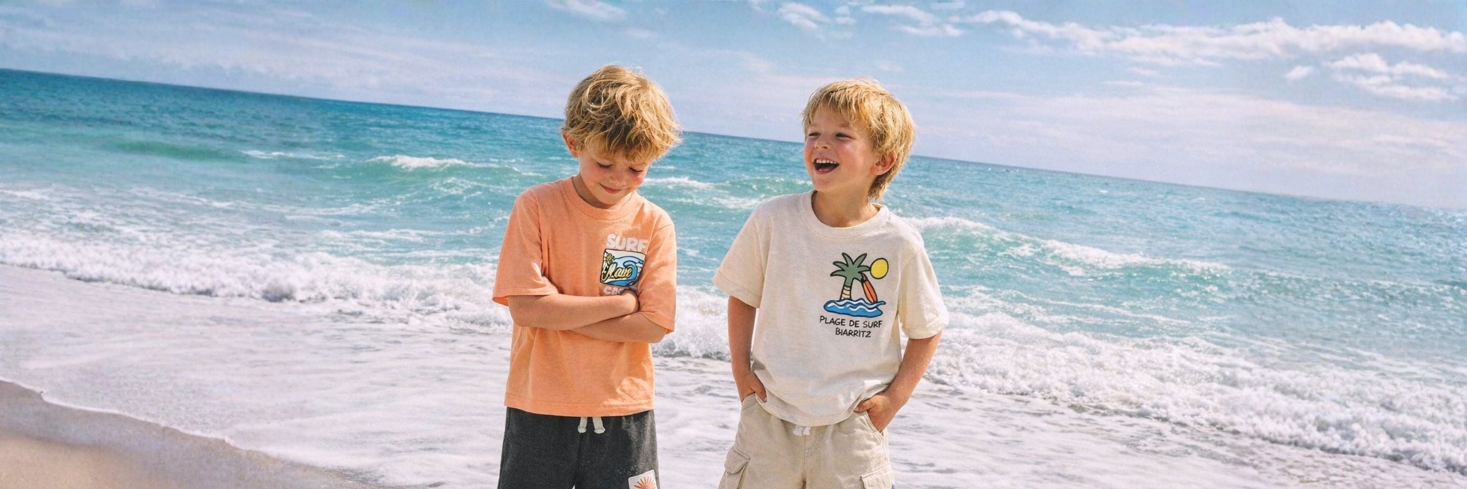 Two boys standing on the beach by the ocean, smiling and laughing, wearing casual T-shirts and shorts with waves in the background.