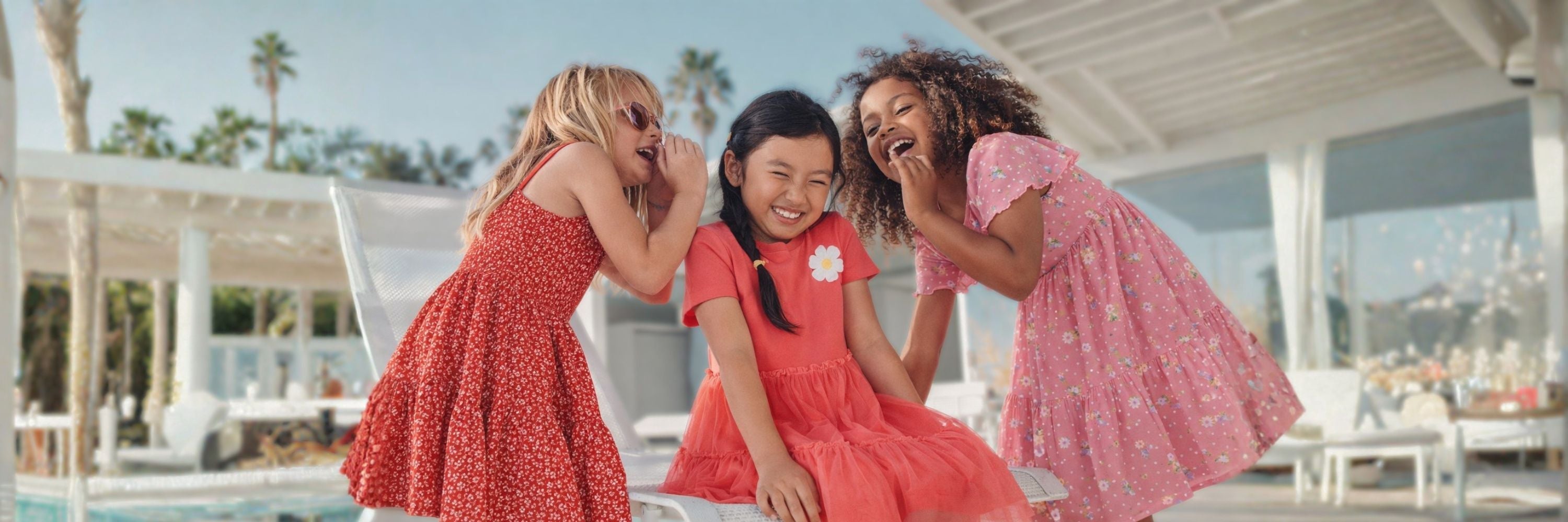 Three girls laughing together outdoors near a pool, wearing colorful summer dresses, sharing a playful moment in a bright sunny setting.