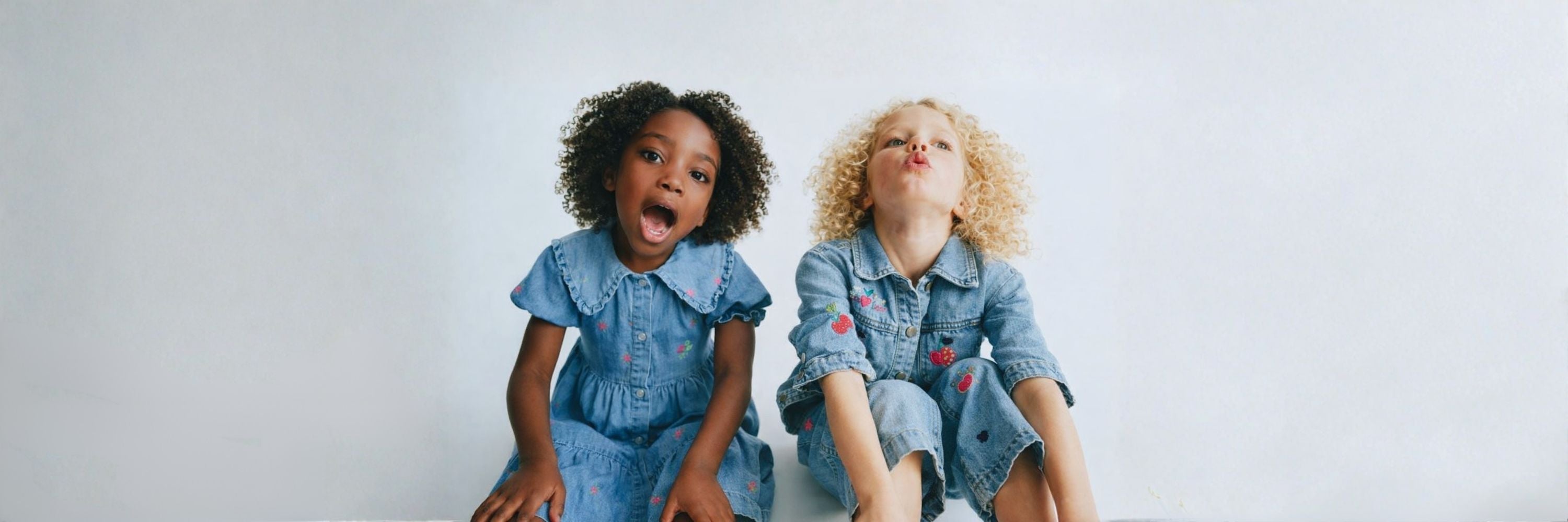 Two girls sitting against a plain background, wearing denim outfits—one in a dress and the other in a matching set—making playful facial expressions.