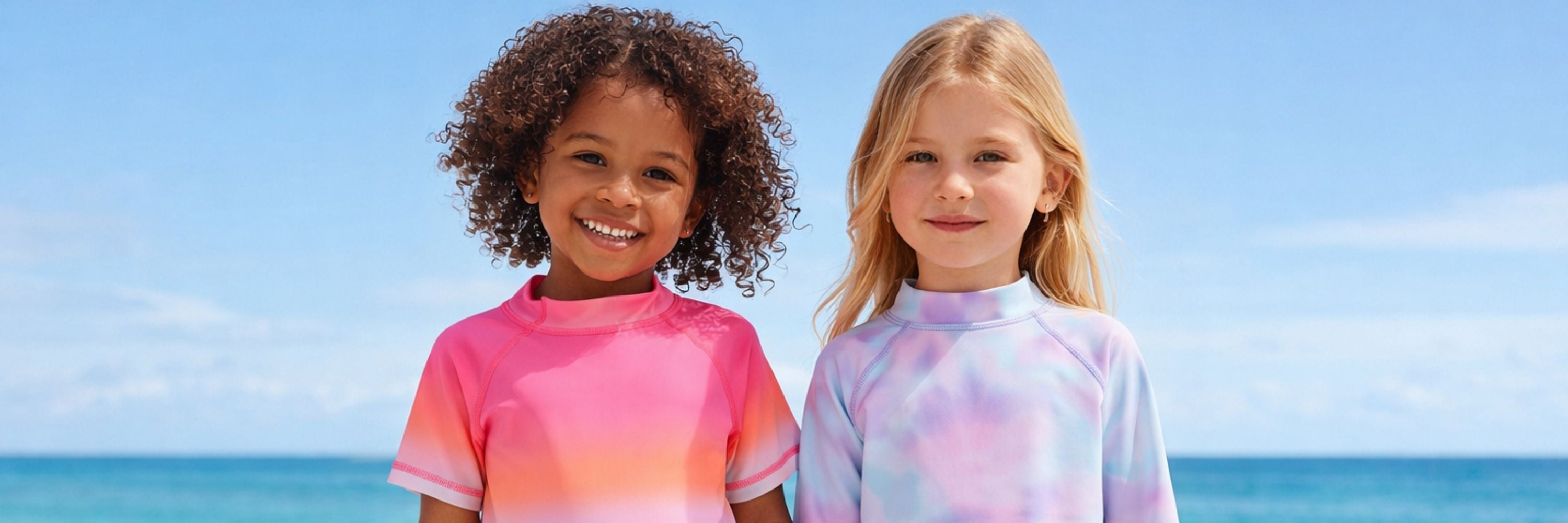 Two smiling girls standing at the beach, wearing colorful long-sleeve swim tops, one in a bright pink gradient and the other in a pastel tie-dye design, with the ocean in the background.