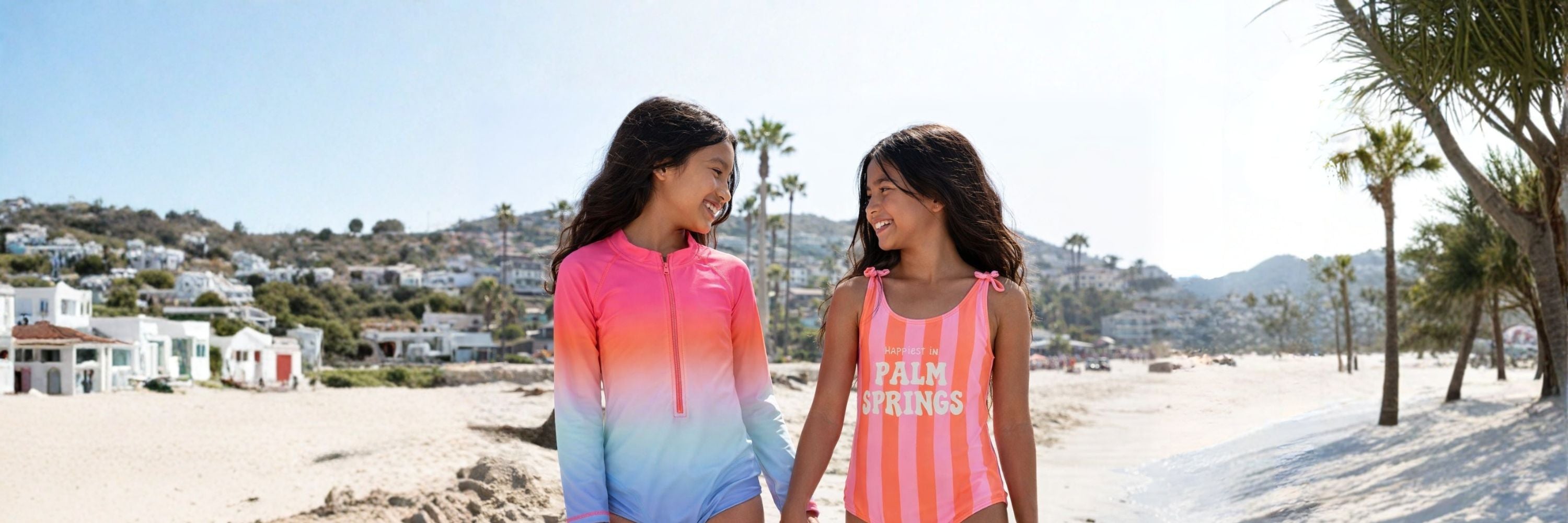 Two girls walking on the beach, smiling at each other, wearing colorful swimwear—one in a gradient long-sleeve suit and the other in a striped “Palm Springs” swimsuit, with palm trees and houses behind.