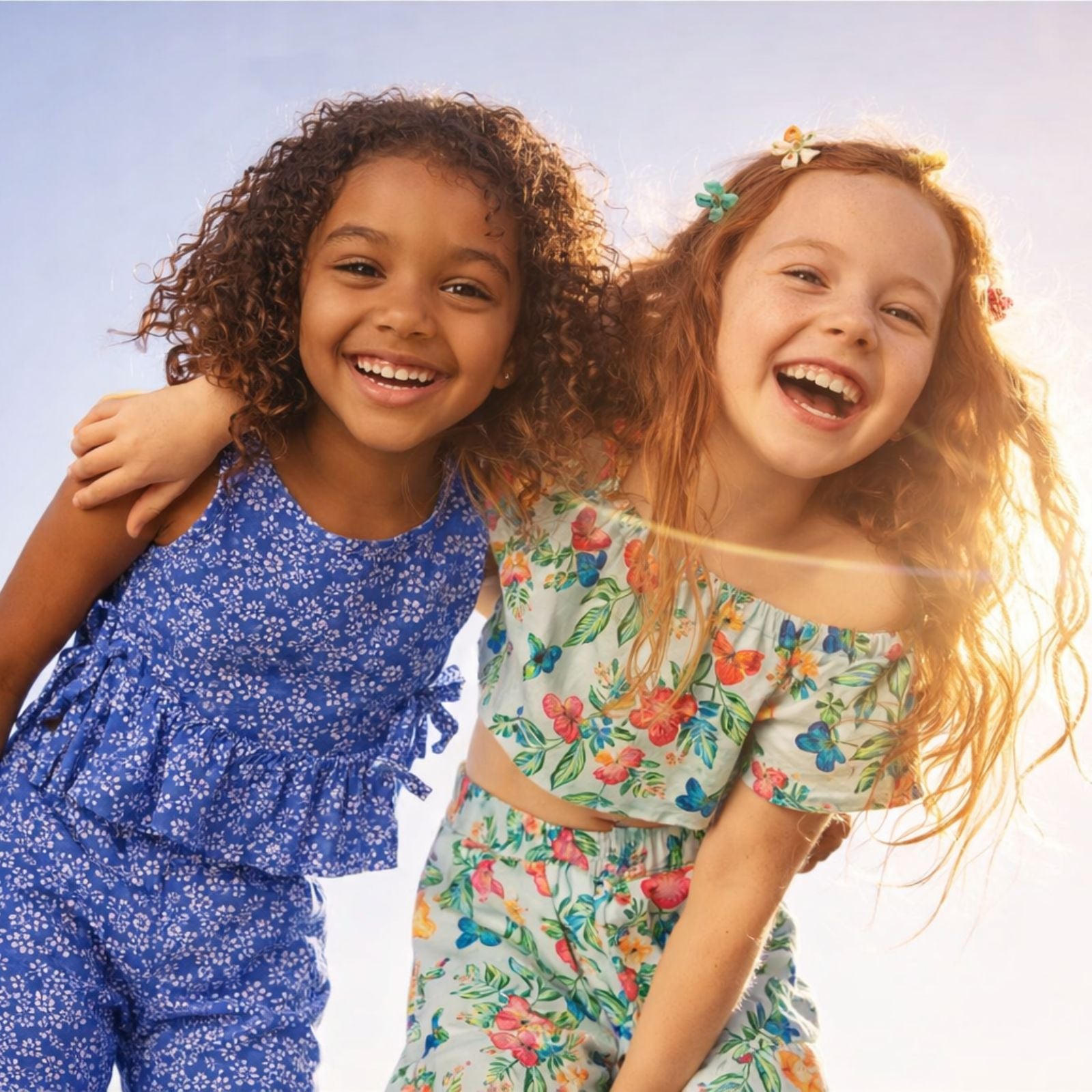 Two young girls in floral outfits smiling against a clear sky.