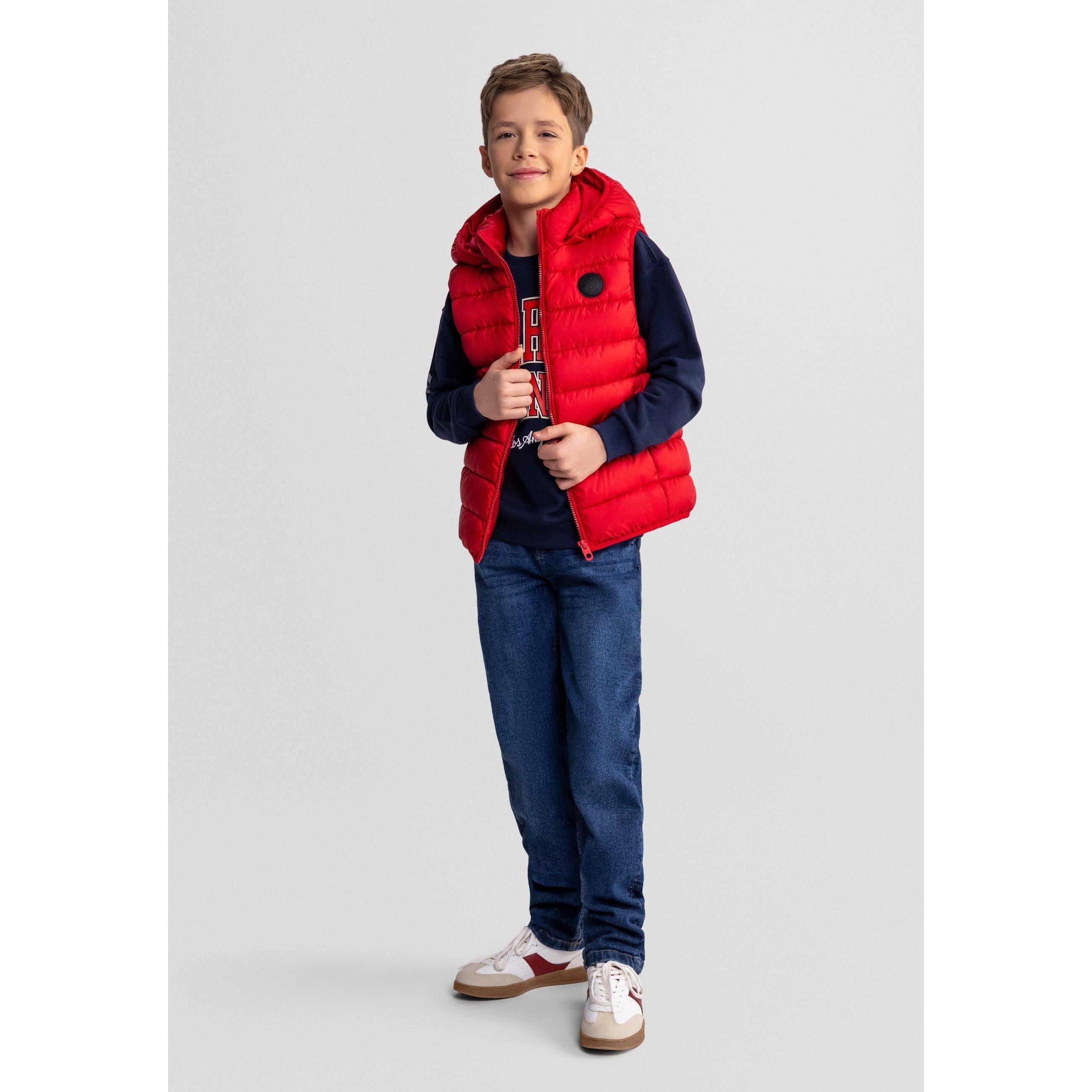 Boy in casual pose wearing red quilted gilet with hood over navy top and blue jeans against studio background