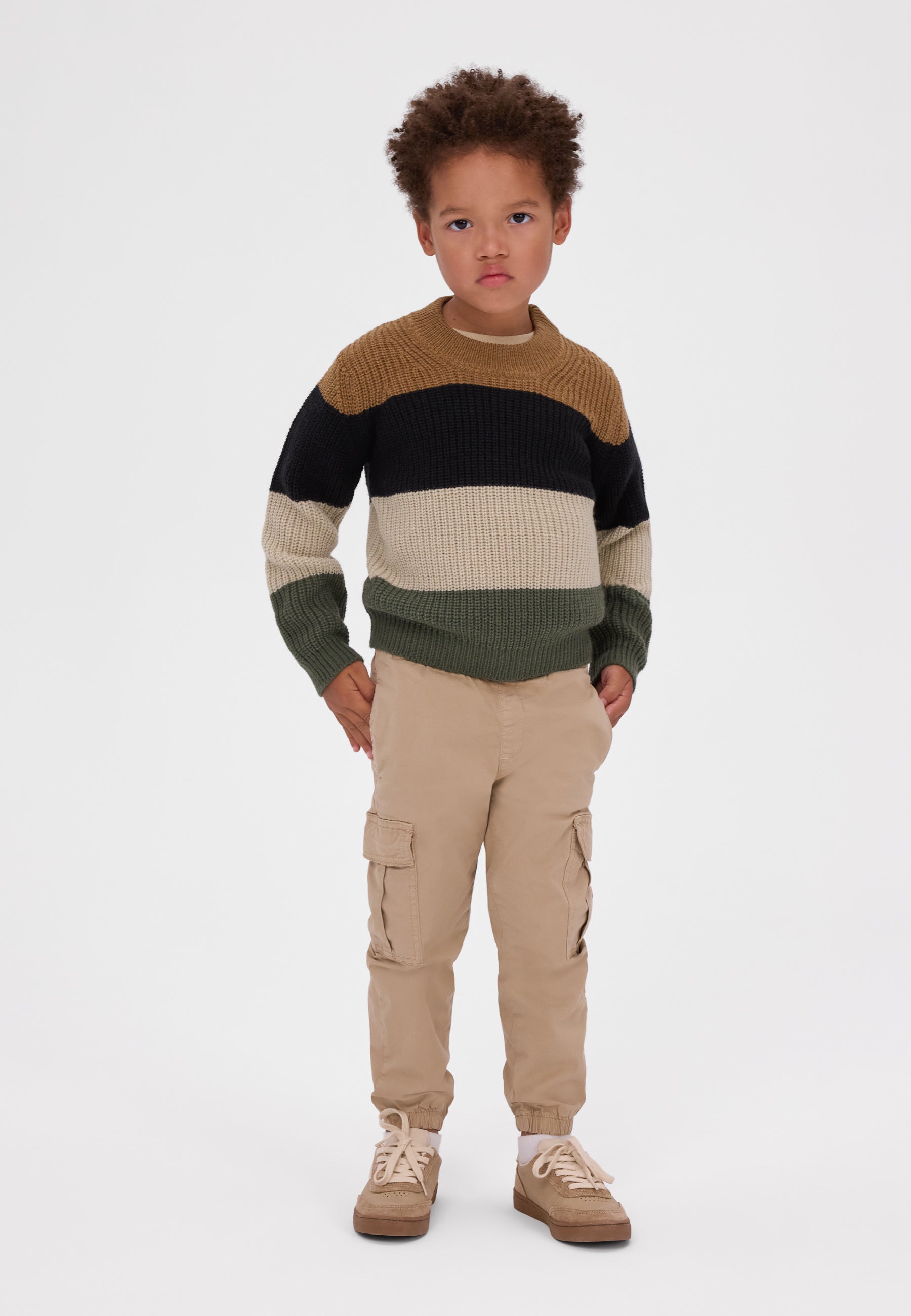 A young child stands against a light gray studio background wearing a color-blocked knit sweater with brown, black, cream, and olive green horizontal stripes, tan cargo pants, and tan sneakers.