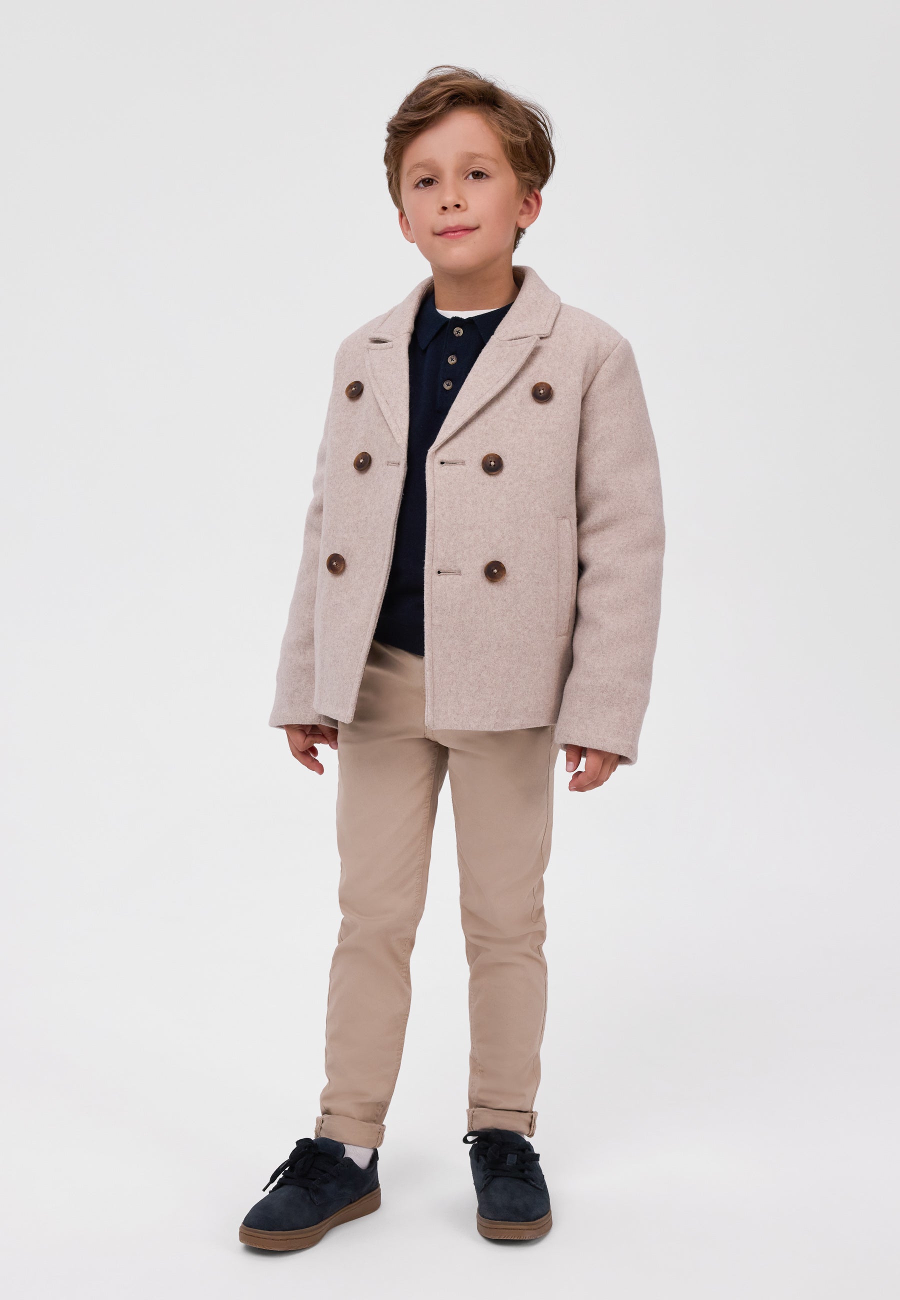 A young boy modeling a light beige double-breasted blazer with brown buttons, paired with beige trousers, a navy polo shirt, and navy sneakers, standing against a plain white studio background.
