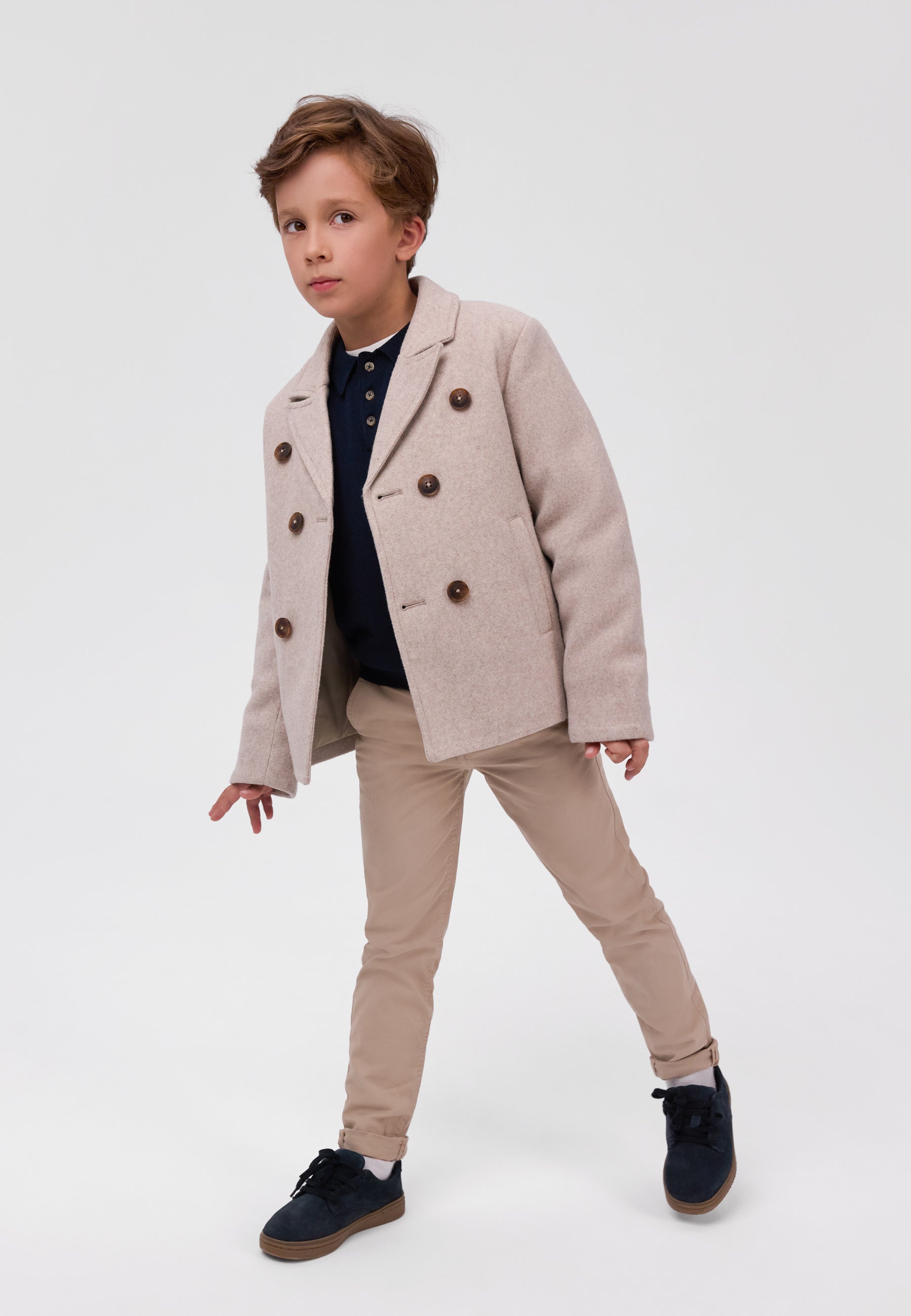 A young boy modeling a light beige double-breasted blazer, beige trousers, a dark navy polo shirt, and navy blue shoes against a plain white studio background.