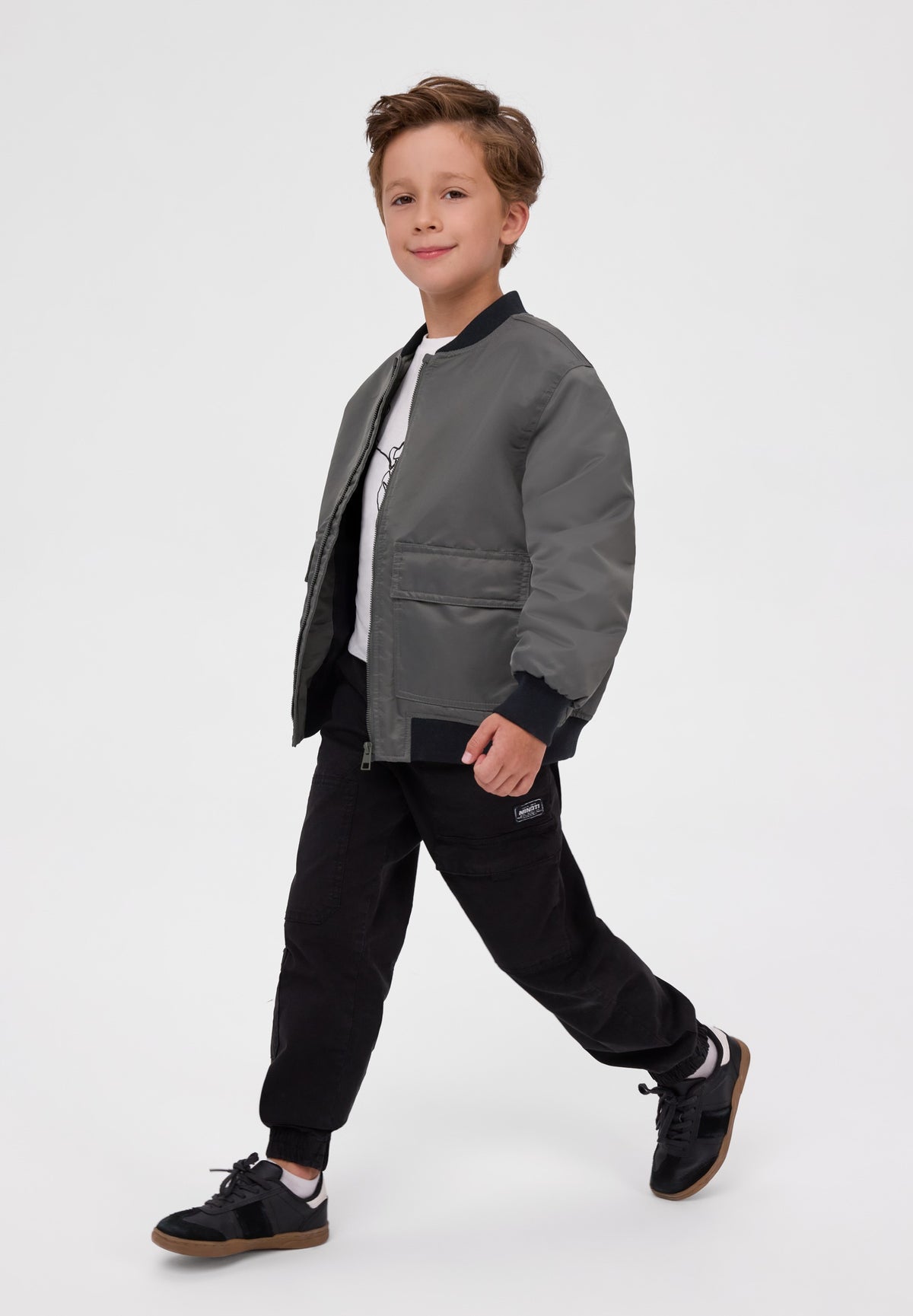 A young boy modeling a dark gray bomber jacket, styled with black joggers, a white t-shirt, and black sneakers, set against a plain light gray studio background.