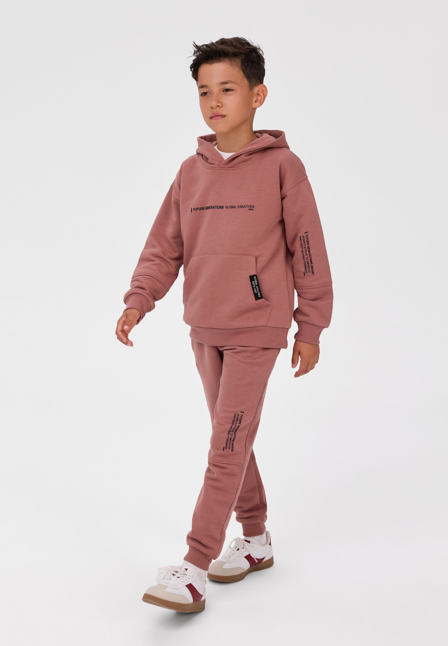 A young boy modeling a dusty rose pink tracksuit, consisting of a pullover hoodie with black text details and matching jogger pants, paired with white sneakers, against a plain light gray studio background.