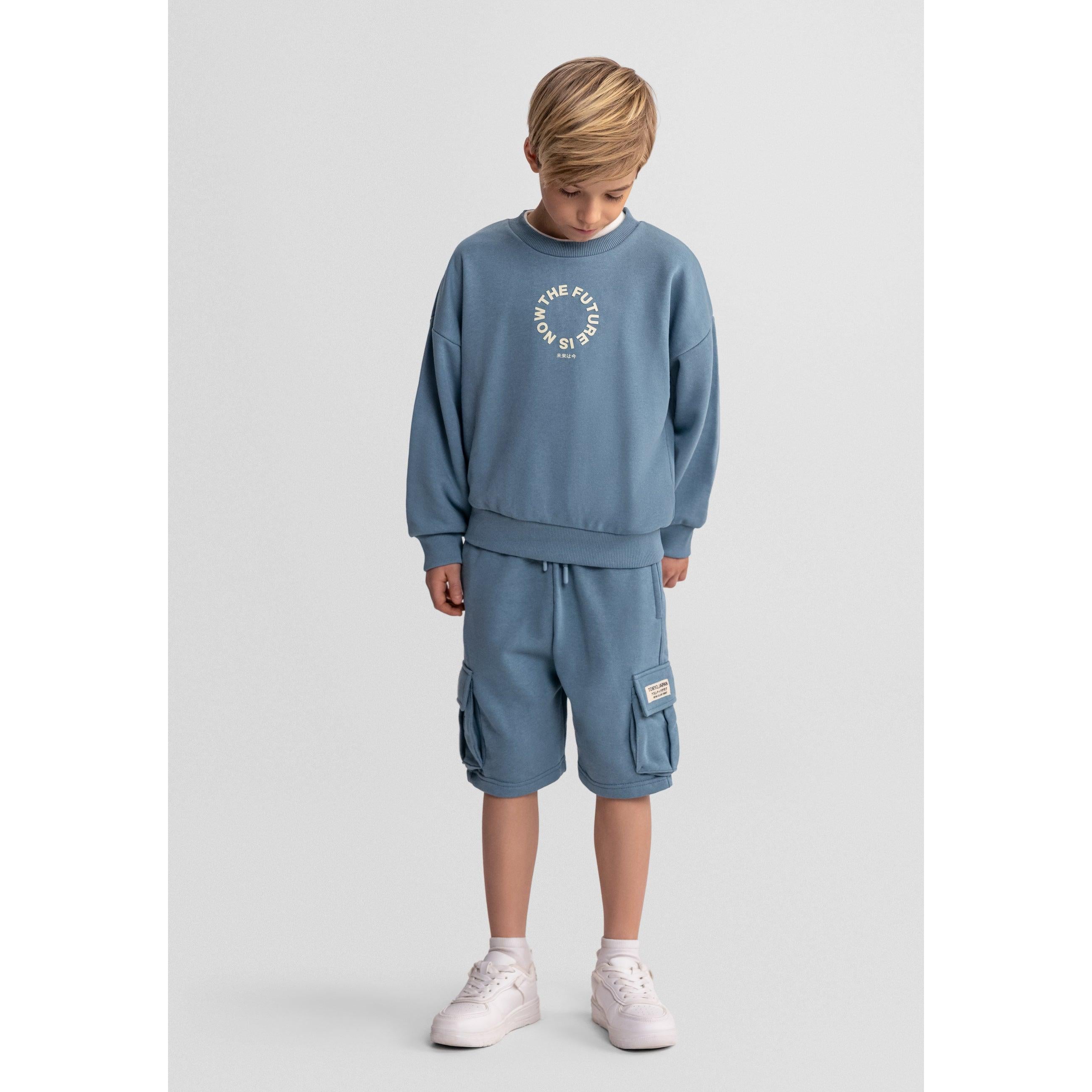 Young blonde boy wearing blue grey sweatshirt and matching cargo shorts set, standing against light grey background